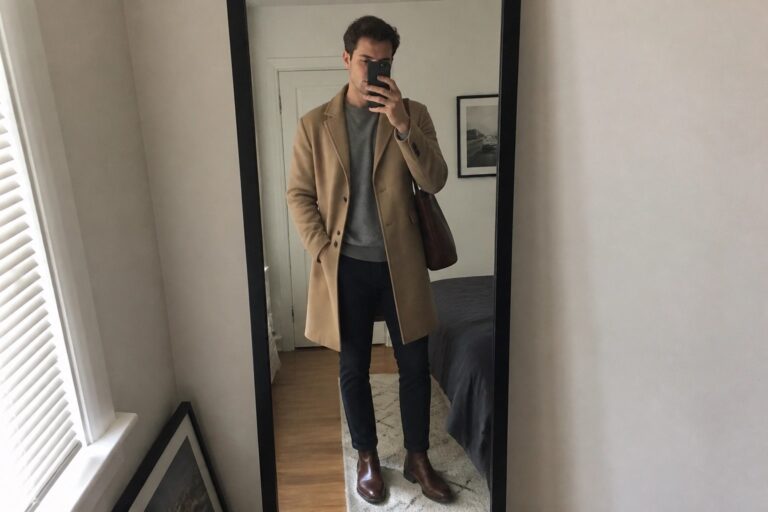 Man in tan overcoat and gray sweater taking bedroom mirror selfie with phone
