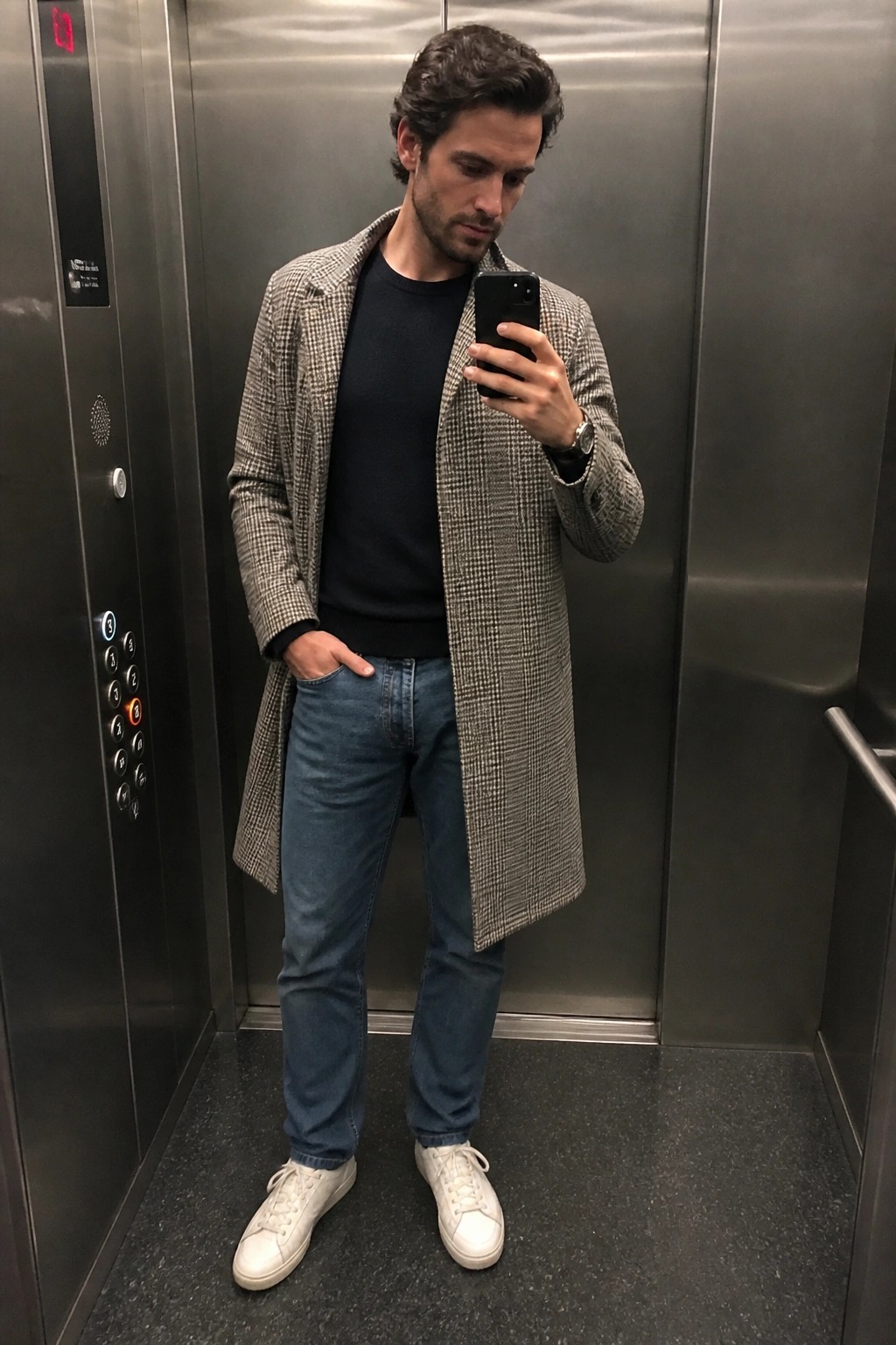 Man standing in elevator wearing long houndstooth overcoat open over navy sweater with blue jeans and white sneakers while holding a phone.