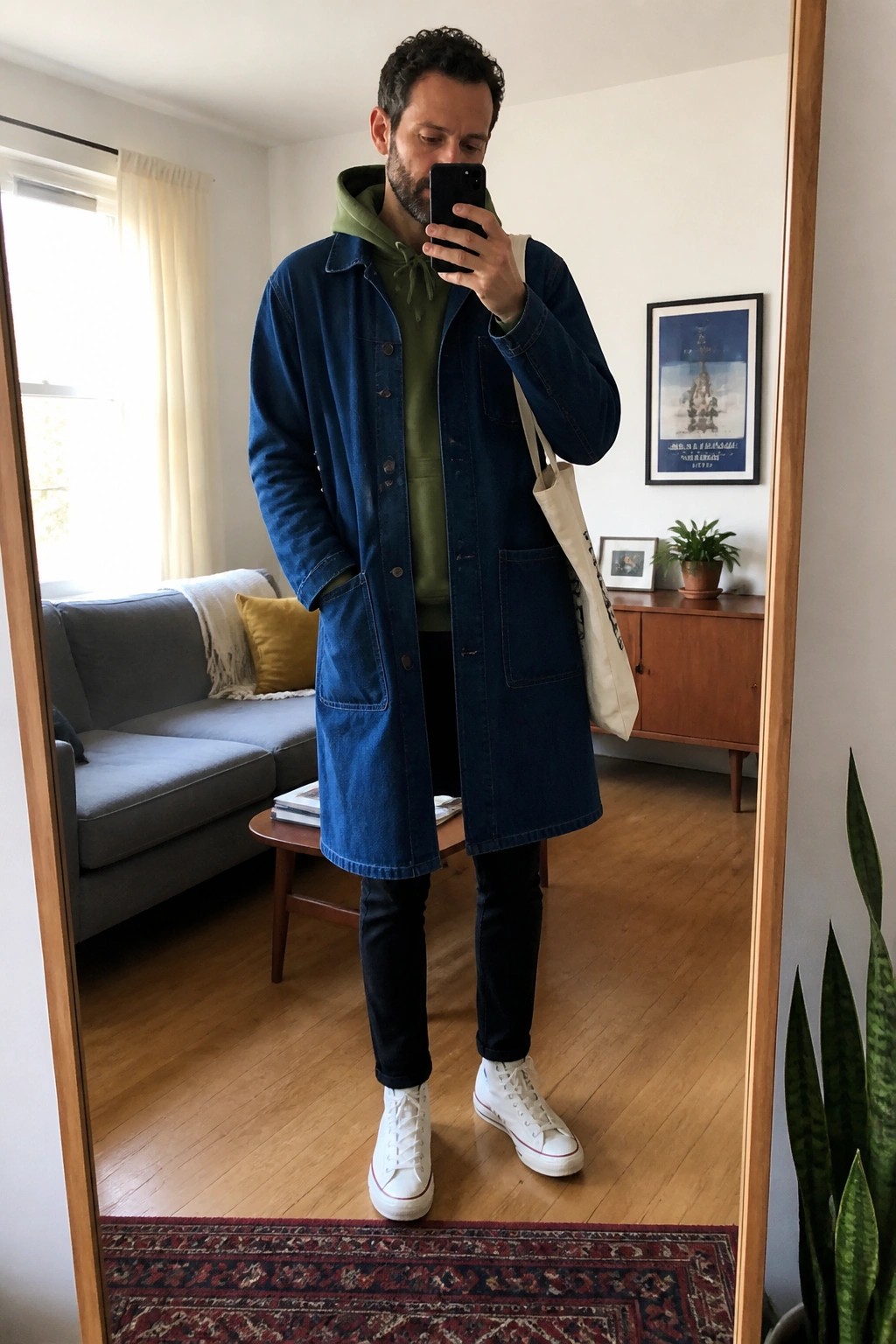 Man in blue denim coat over green hoodie taking mirror selfie in living room