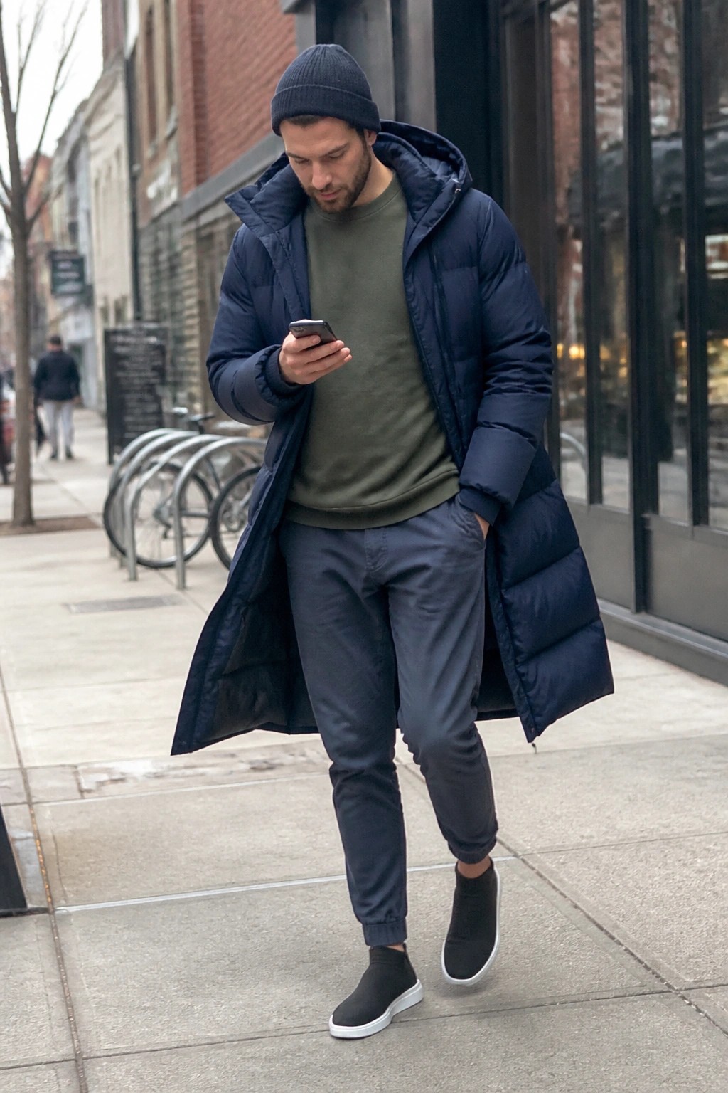 Man in navy puffer coat and beanie checks smartphone on urban sidewalk