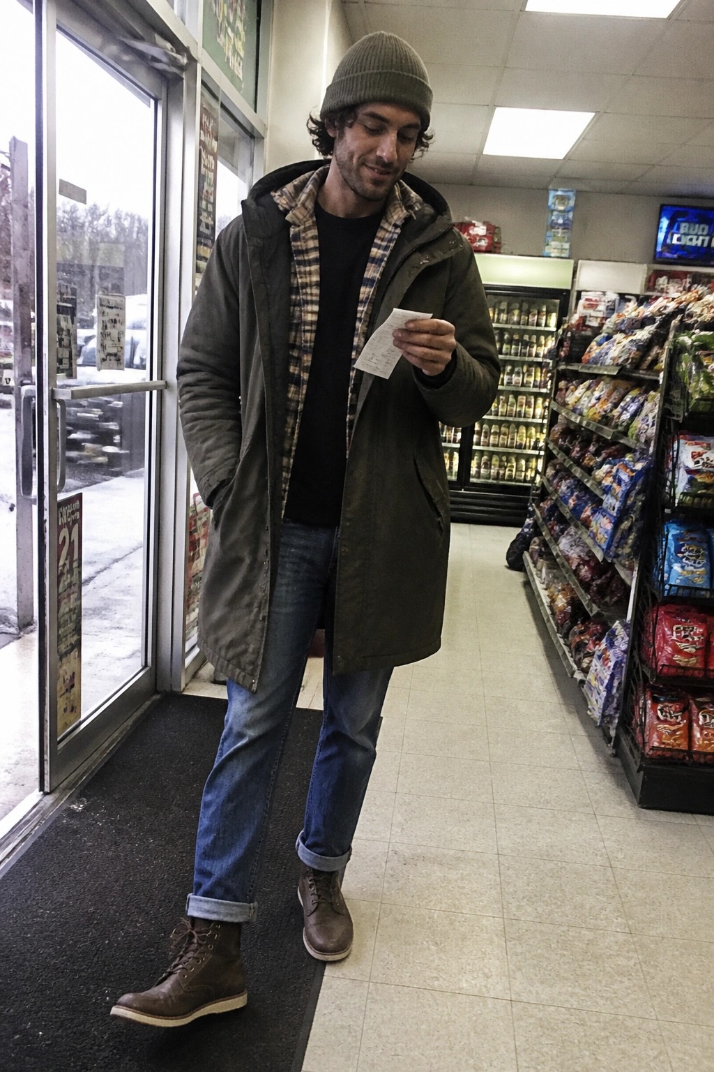 Man in green parka and beanie examines receipt inside convenience store