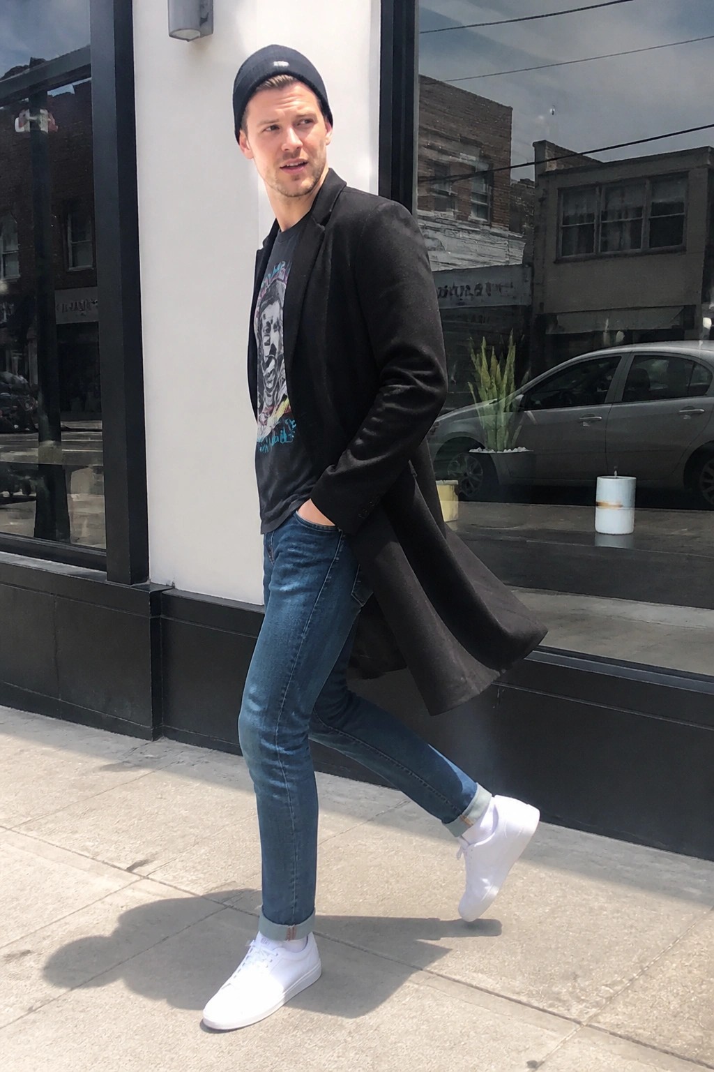 Man in black blazer over graphic tee, blue jeans, and white sneakers on sidewalk