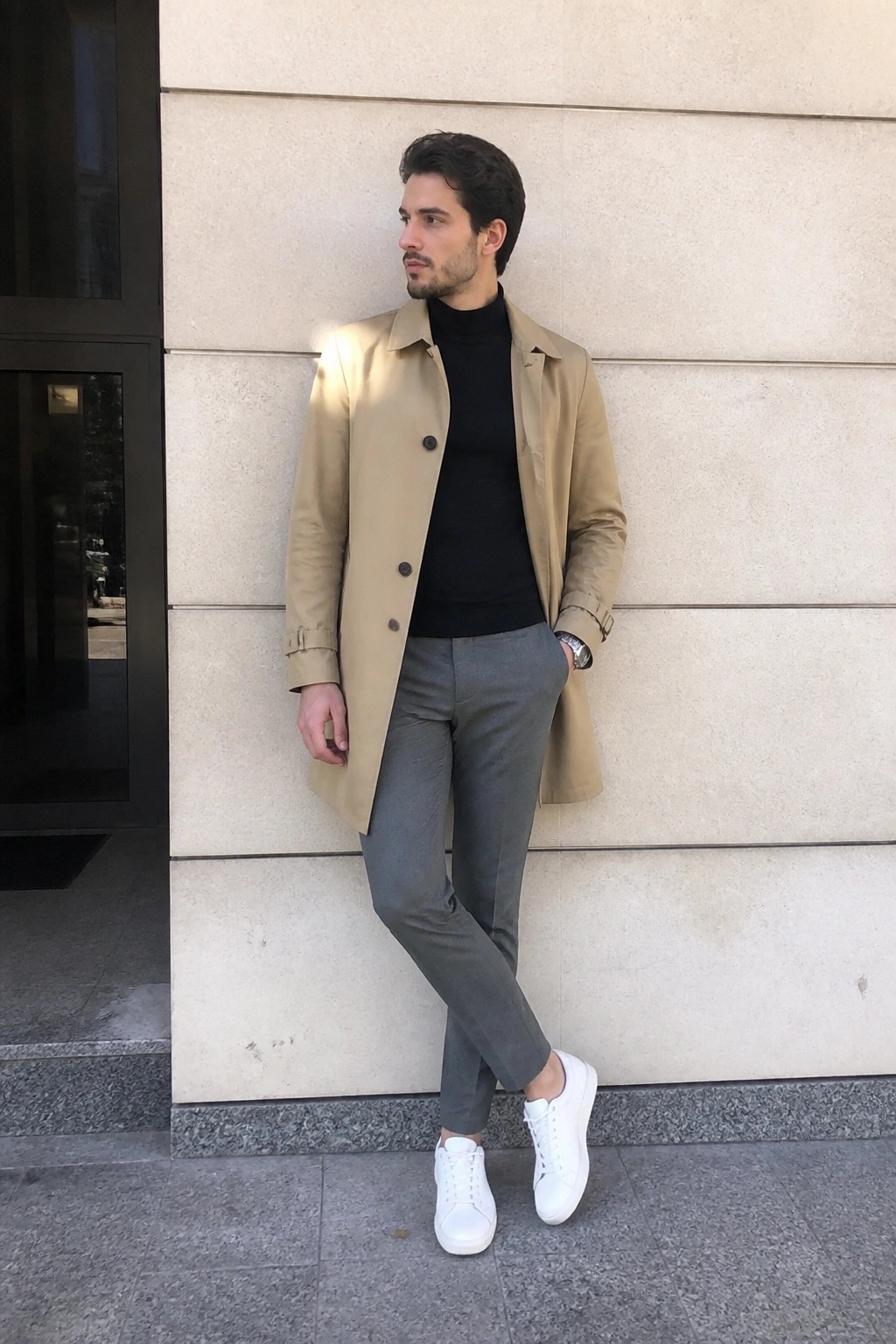 Man in beige trench coat, black turtleneck, gray pants and white sneakers by stone wall
