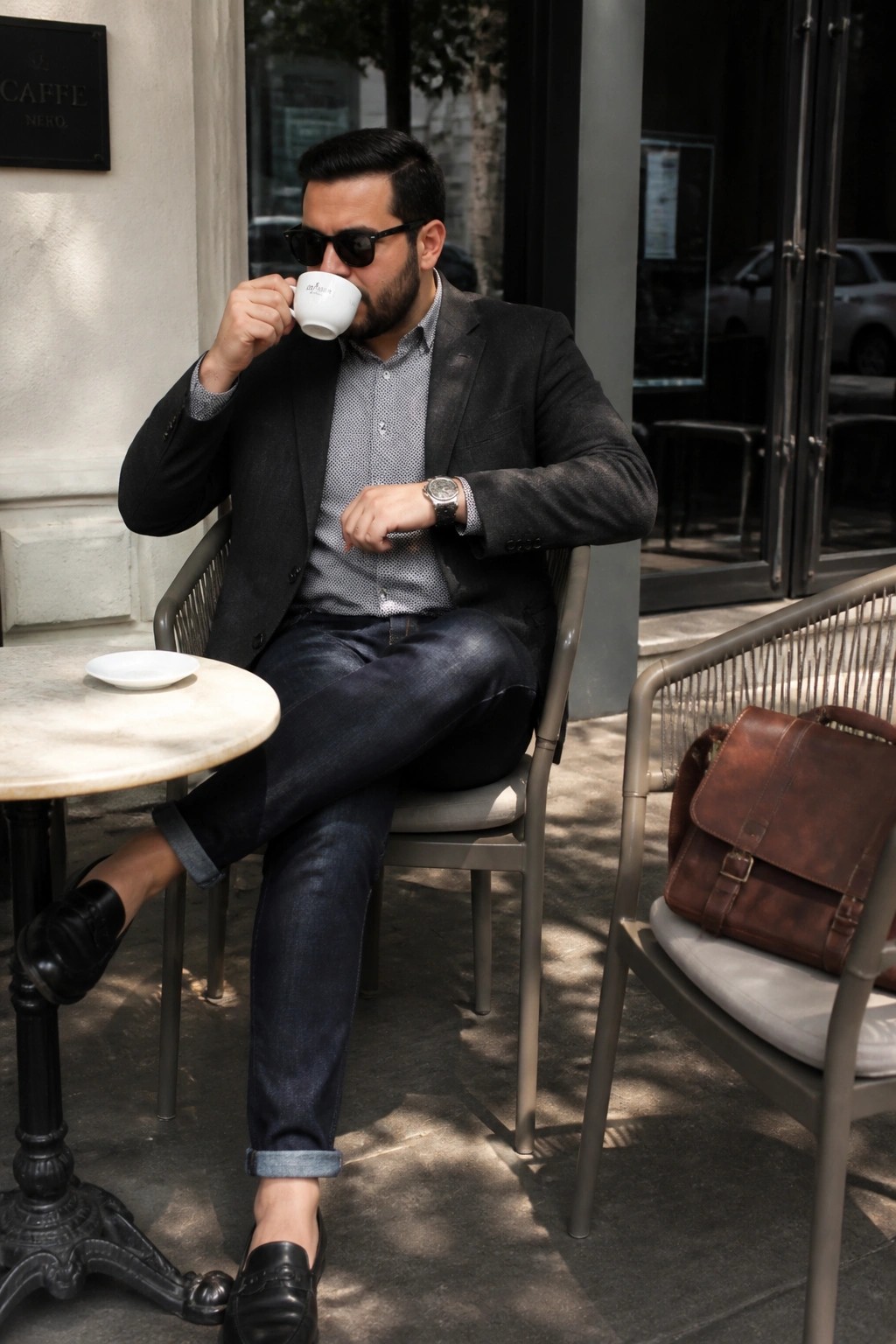 Bearded man in blazer sips coffee while checking watch beside leather bag at cafe