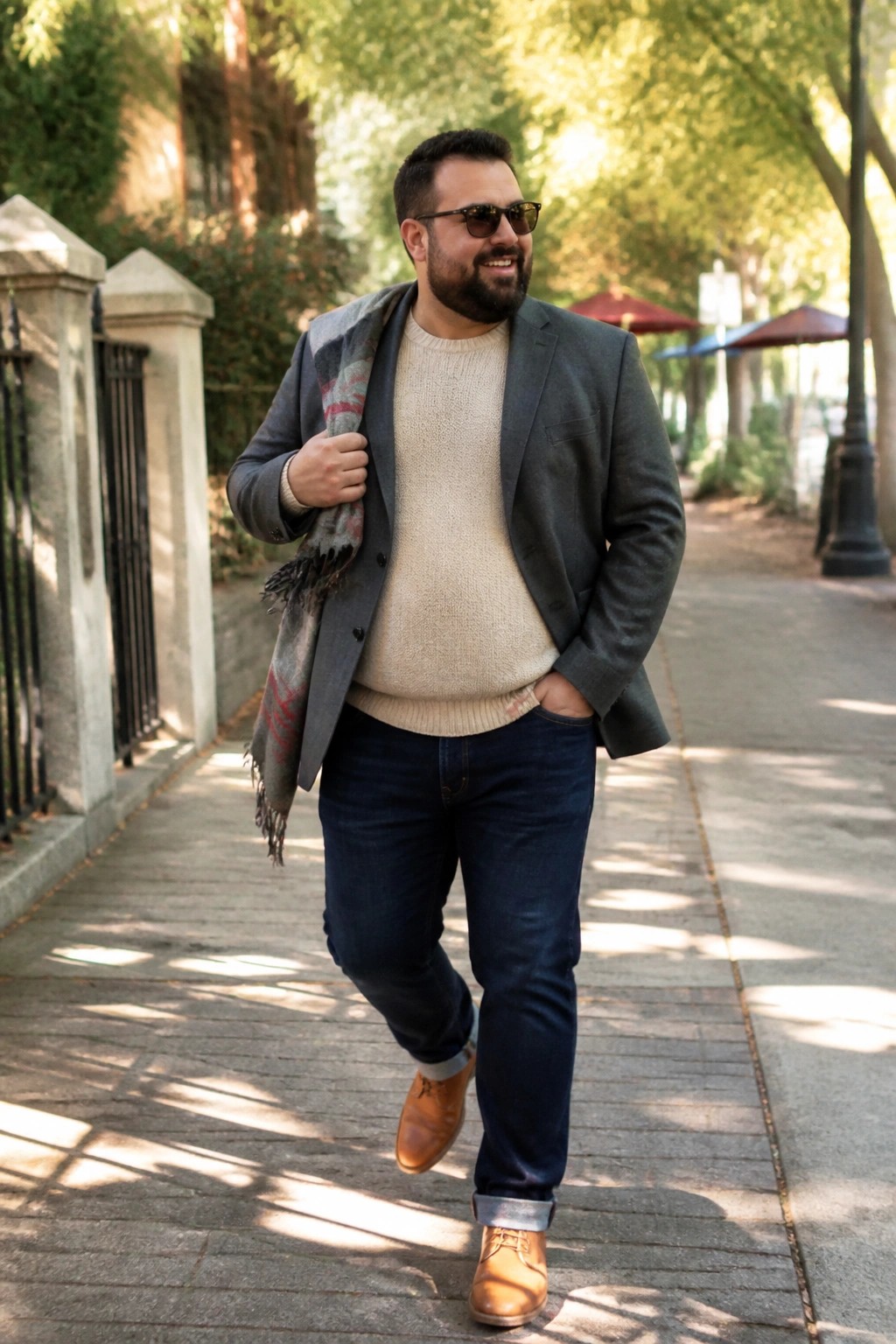Bearded man in gray blazer, cream sweater, and blue jeans walks smiling on sunny path