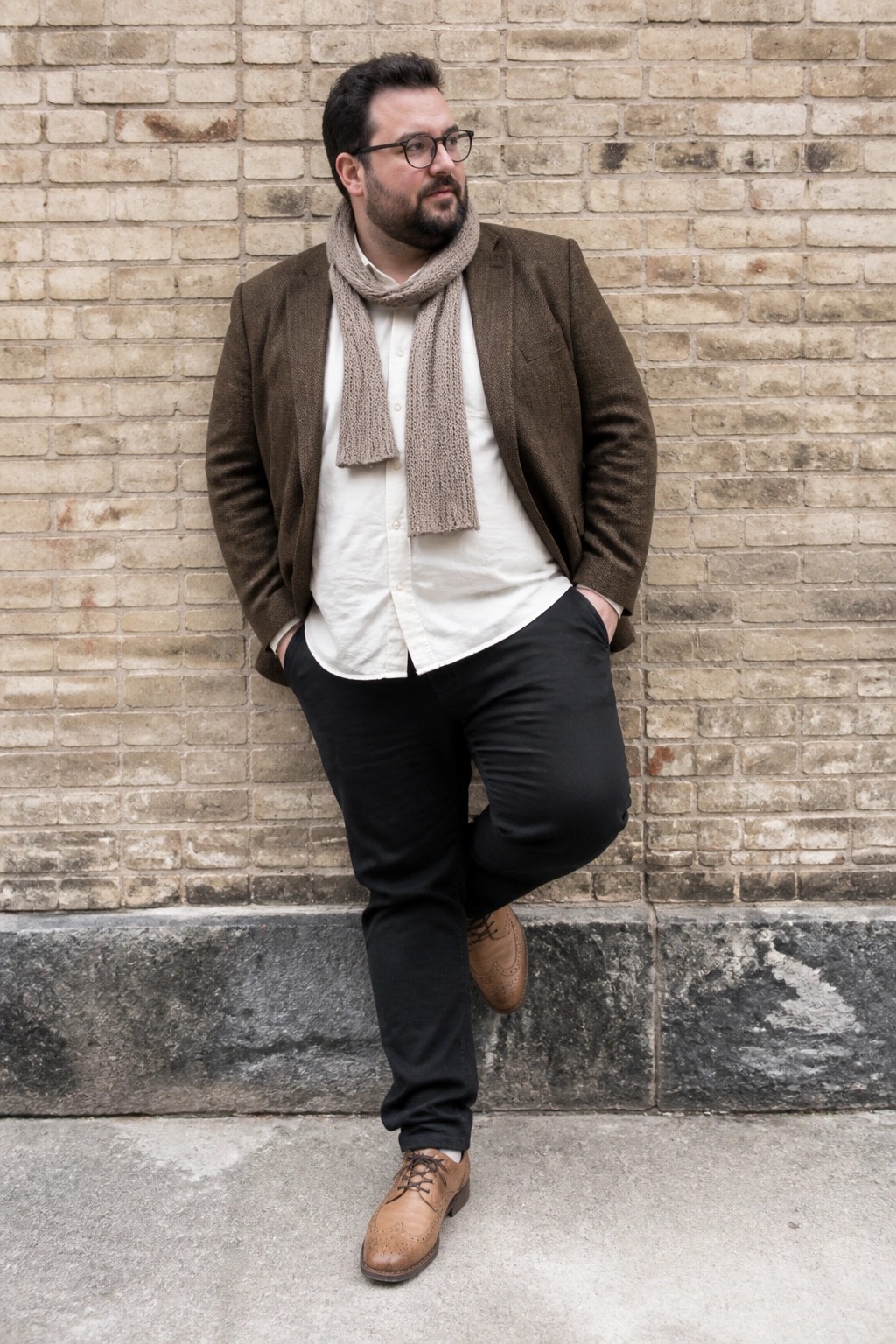 Bearded man in brown blazer and knit scarf posing against brick wall with one leg raised.