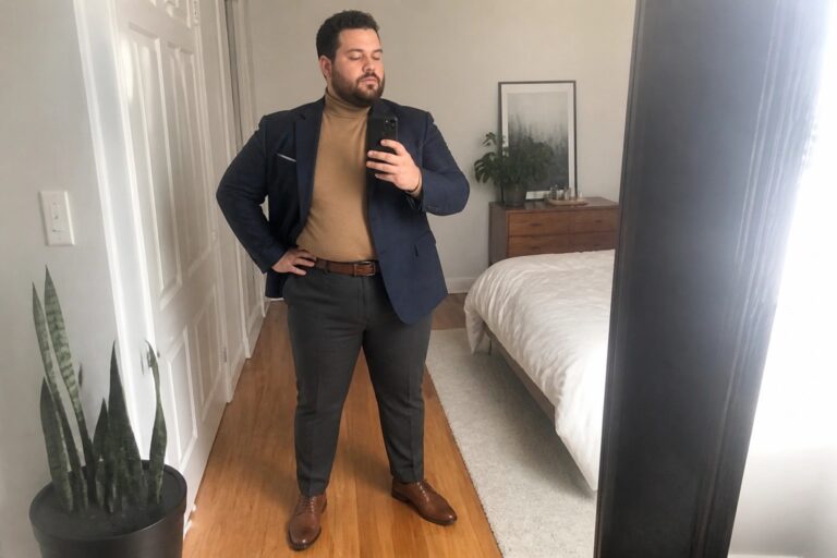 Man in navy blazer and tan turtleneck taking mirror selfie in bedroom