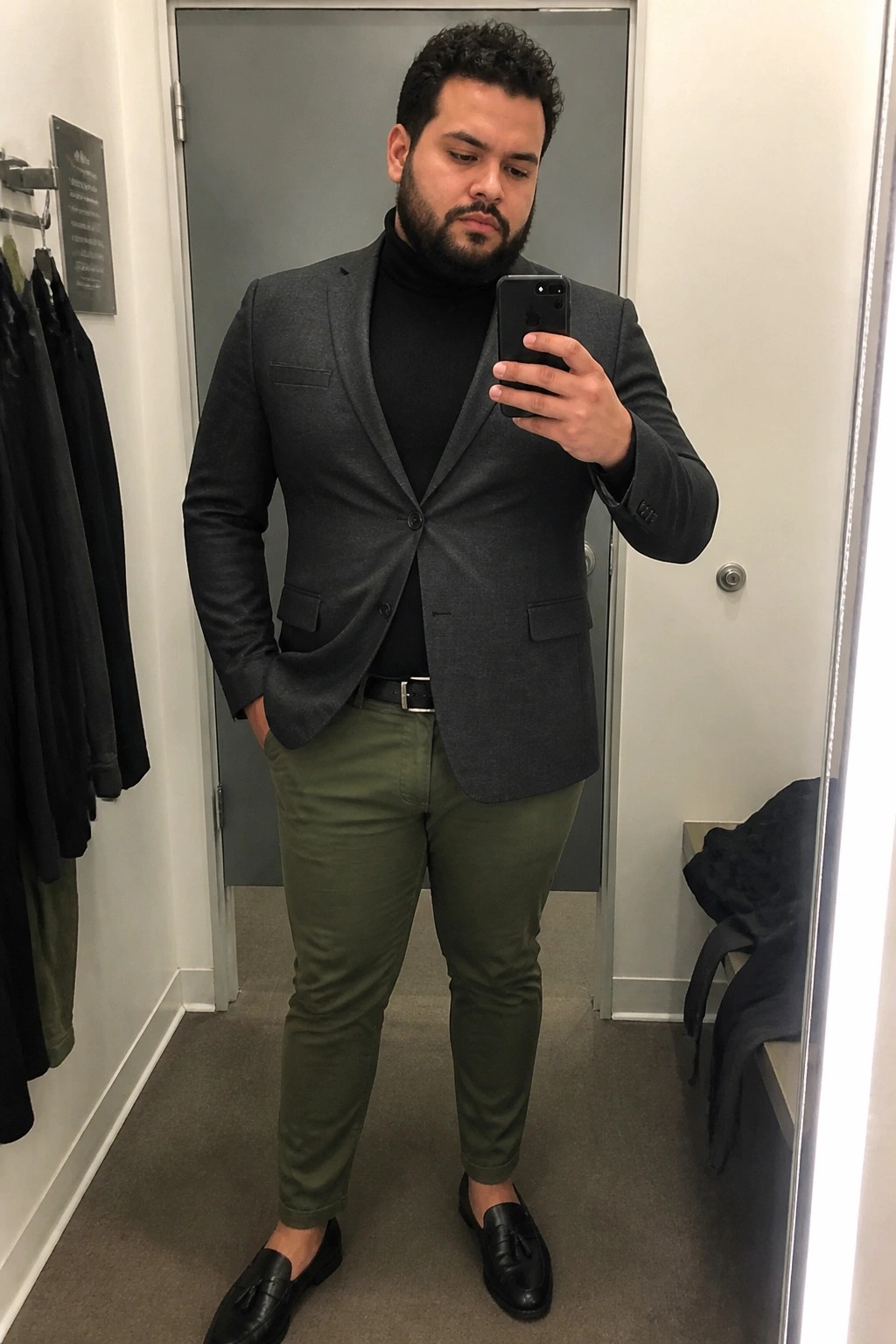 Man in gray blazer over black turtleneck with olive pants taking mirror selfie
