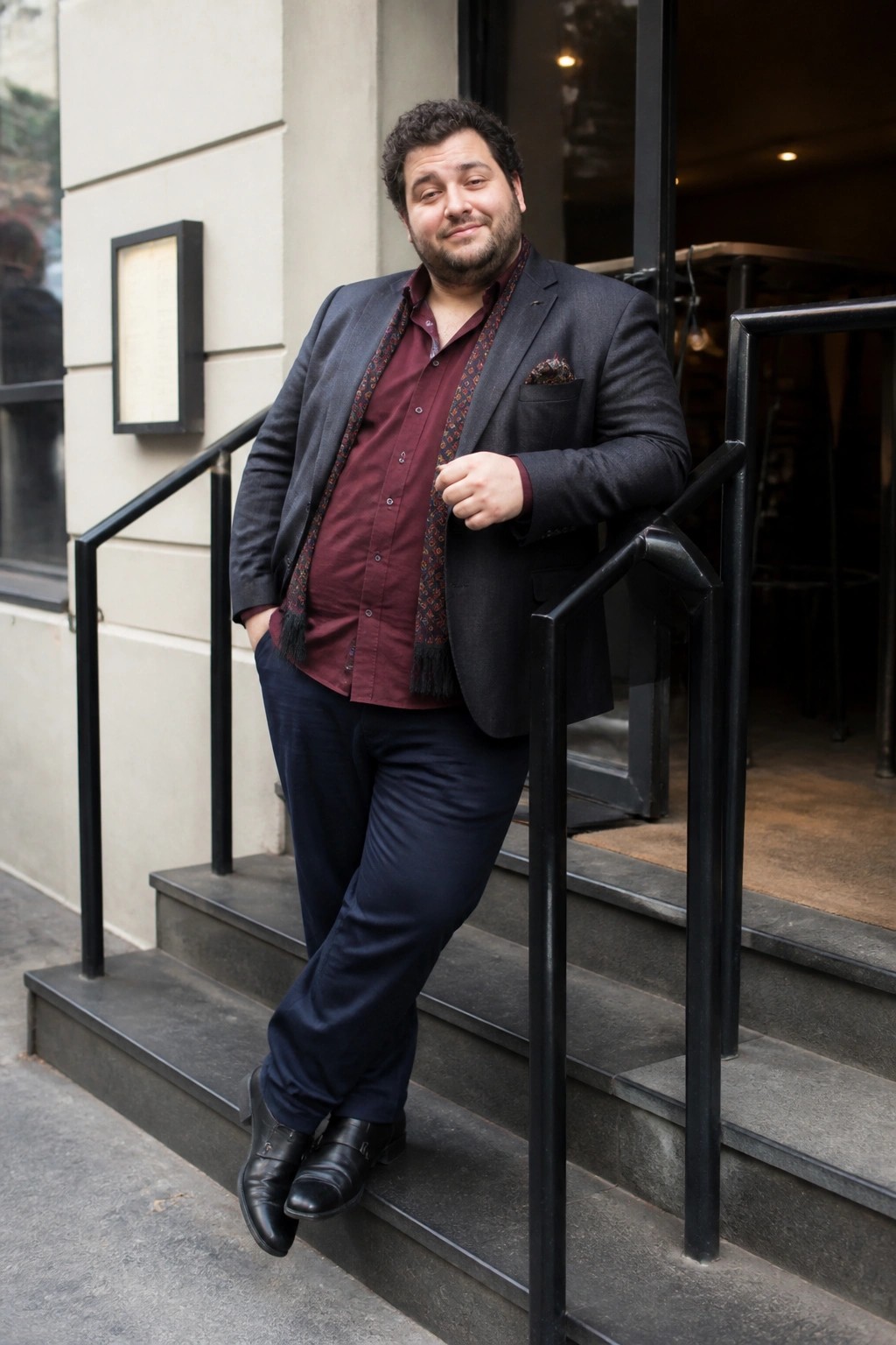 Curly-haired man in gray blazer and maroon shirt leans on stair railing.