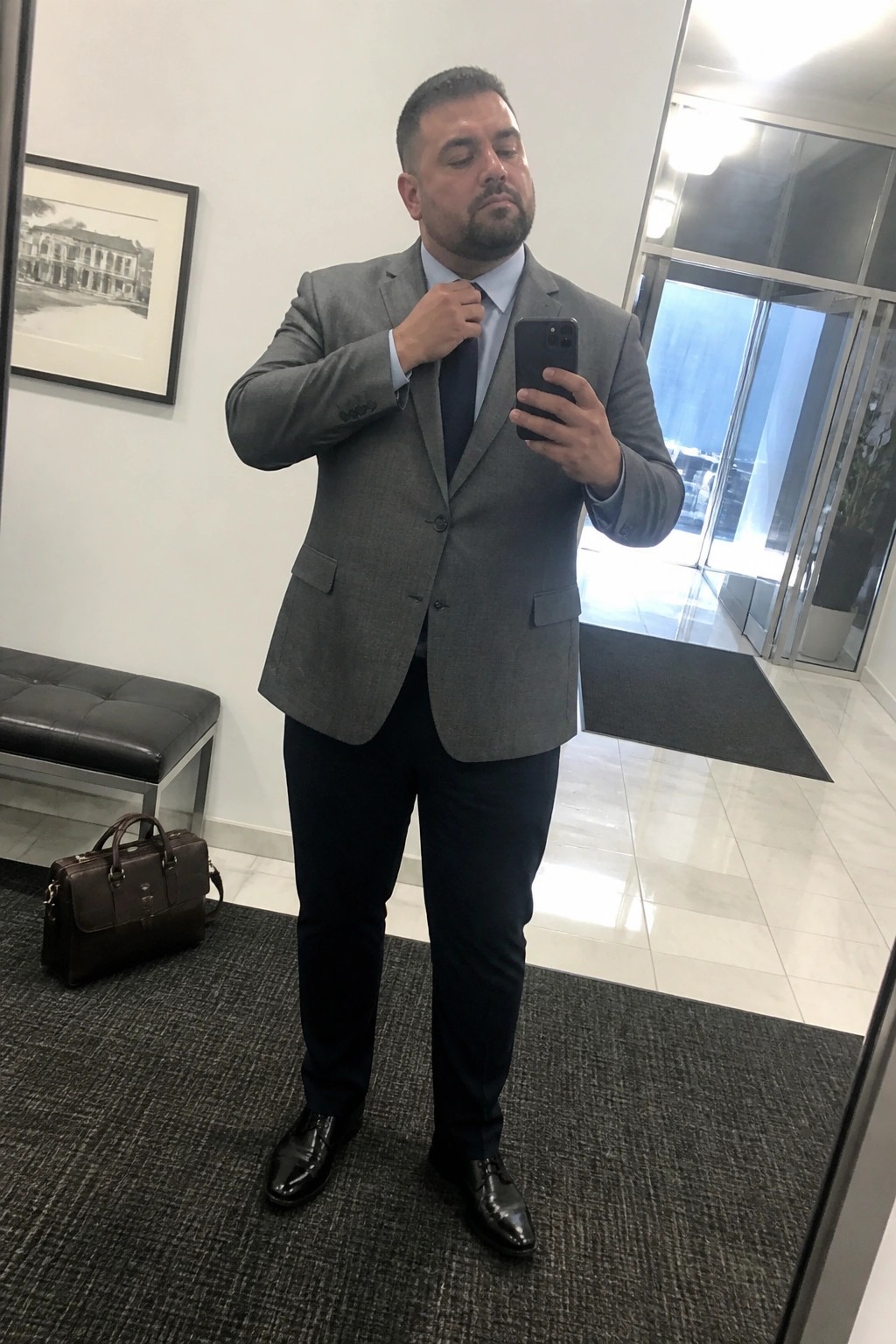 Man in gray suit adjusts navy tie while taking mirror selfie with phone