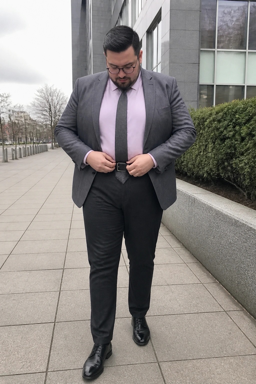Man in gray suit with pink shirt adjusts belt on urban sidewalk