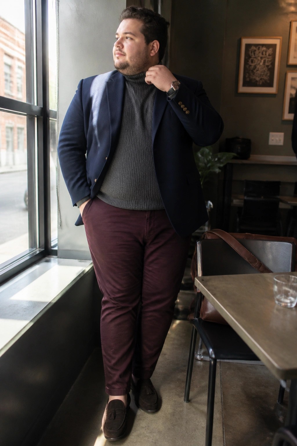 Man in navy blazer, gray turtleneck, maroon pants adjusting collar in cafe