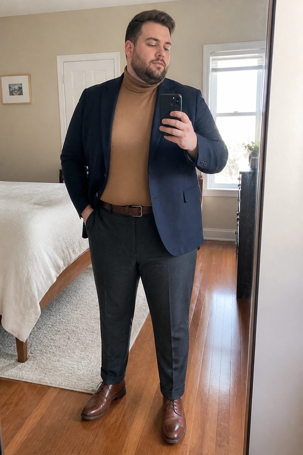 Man in navy blazer and tan turtleneck taking mirror selfie in bedroom