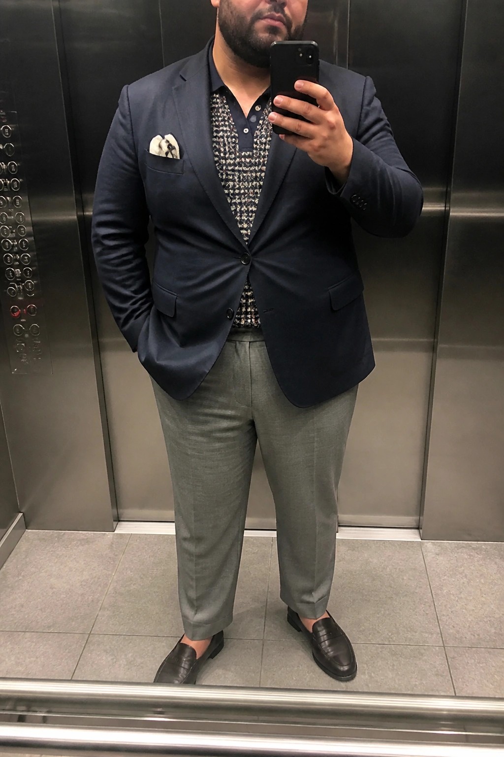 Man in navy blazer over patterned shirt taking mirror selfie in elevator