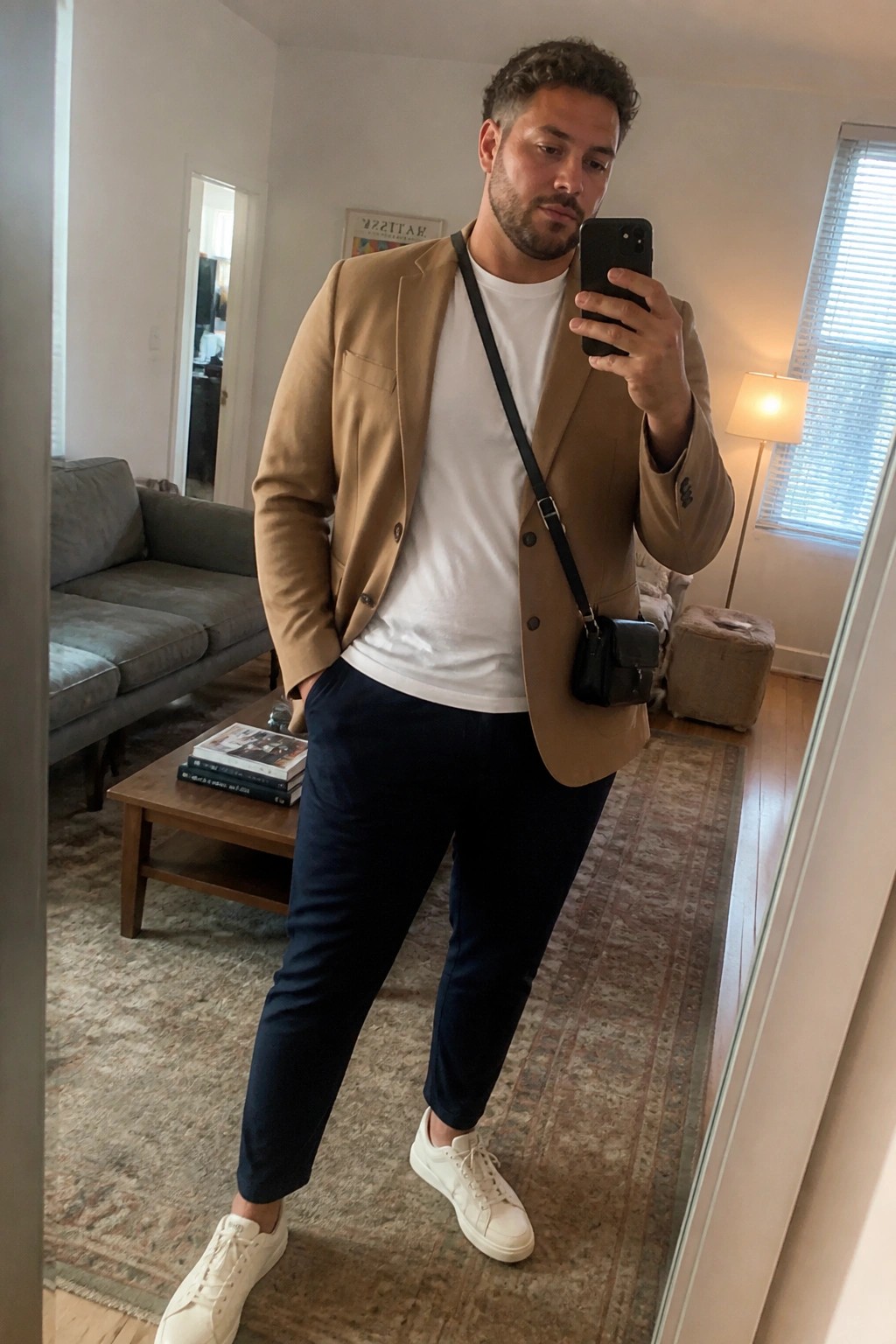 Man in tan blazer, white tee, navy pants taking mirror selfie in living room