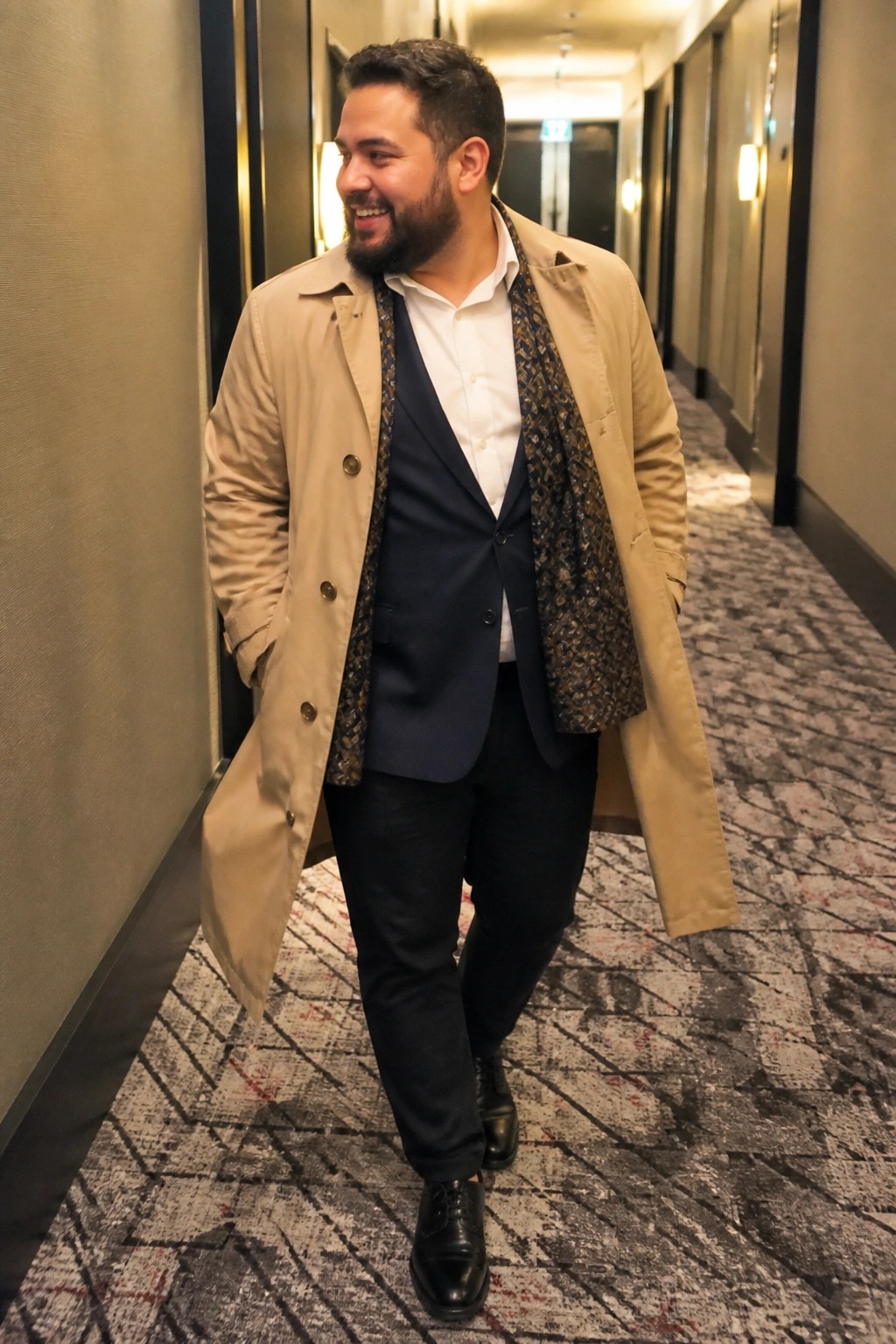 Smiling bearded man in beige trench coat and navy vest walks in hallway.