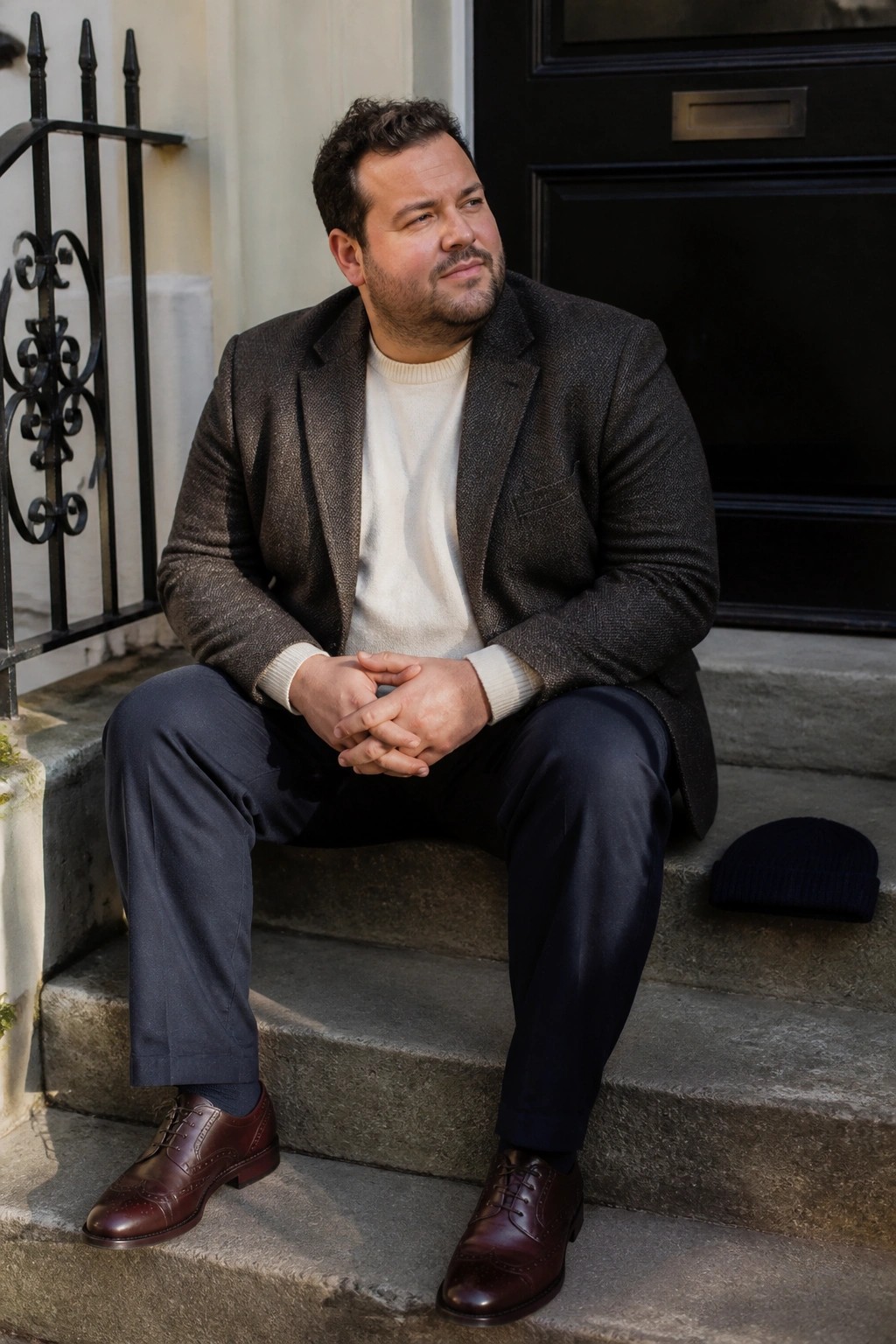 Man in brown tweed blazer and cream sweater sits on stone steps beside black beanie