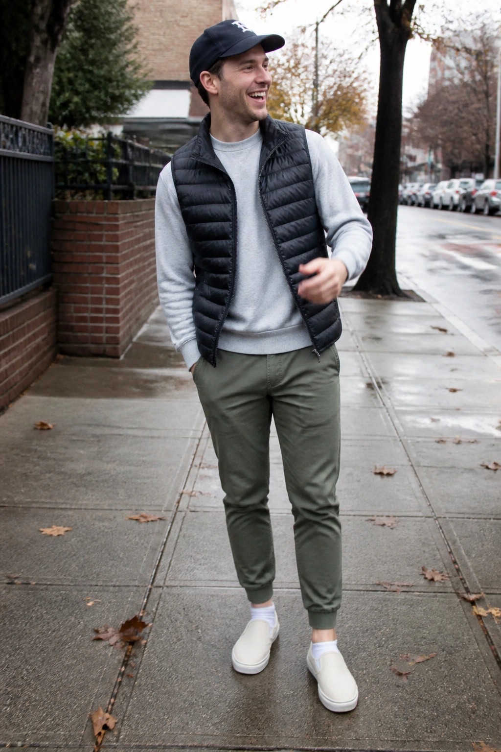 Man in black puffer vest over gray sweatshirt with olive pants on wet sidewalk