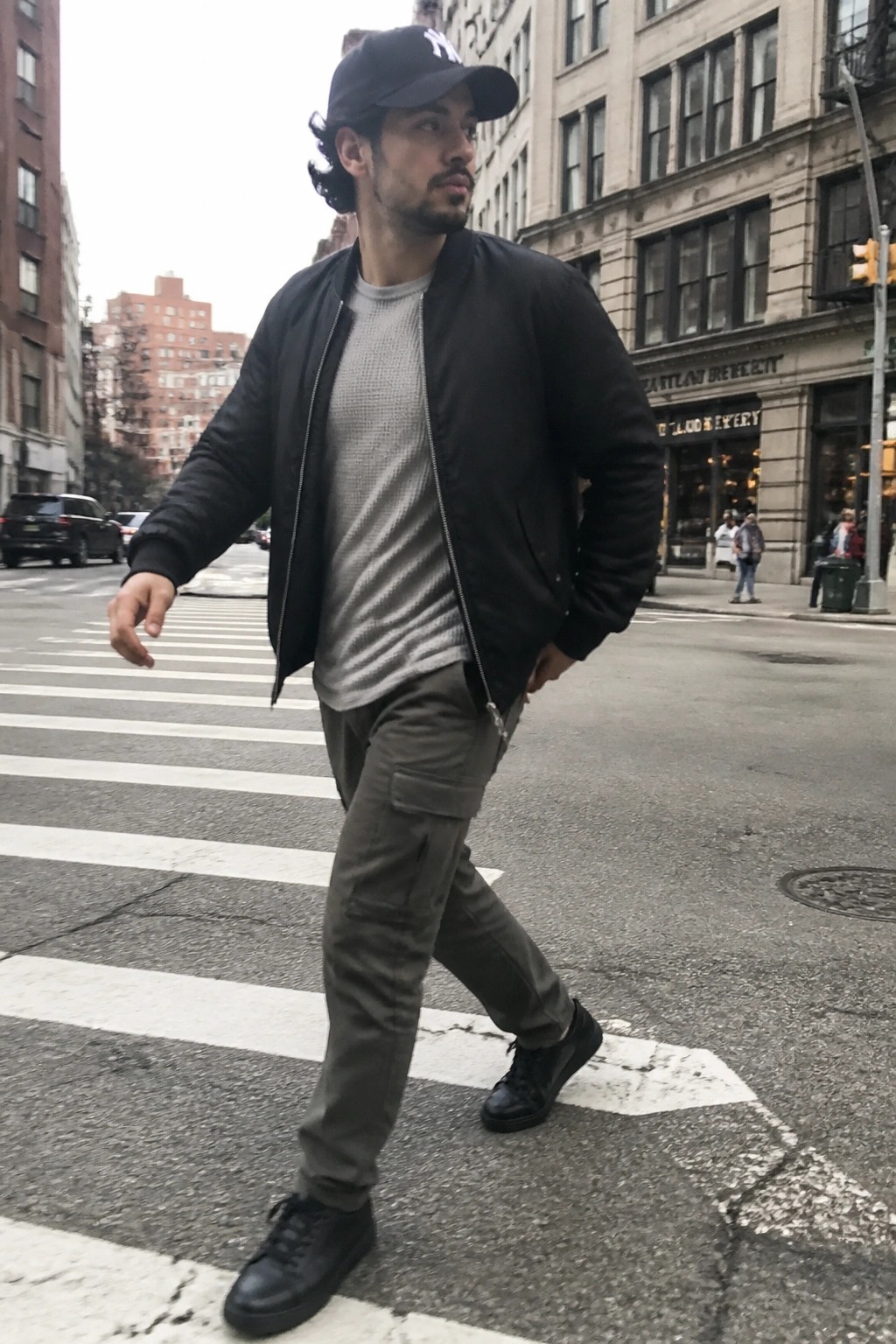 Man in black NY cap, bomber jacket, and cargo pants crossing city street