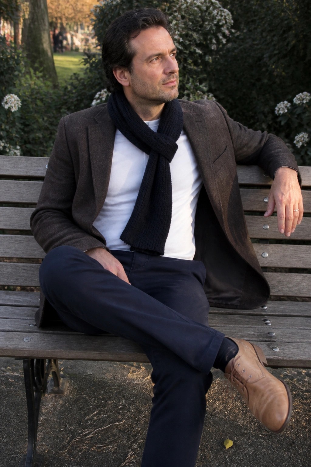Man seated on wooden bench wearing brown blazer over white t-shirt with black knit scarf, dark blue trousers, and brown leather shoes in casual pose.