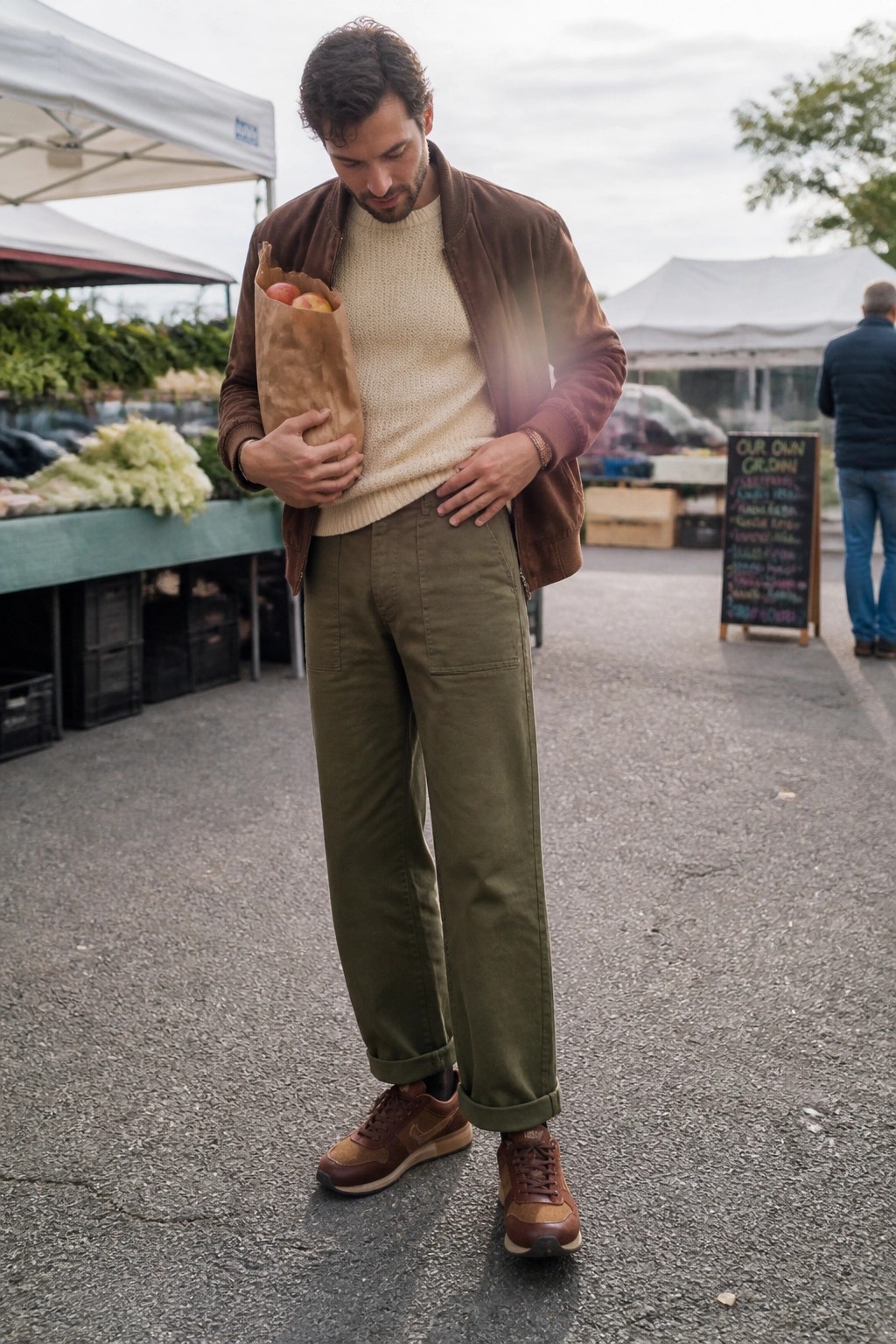 Man in brown jacket and olive pants holding paper bag of apples at market.