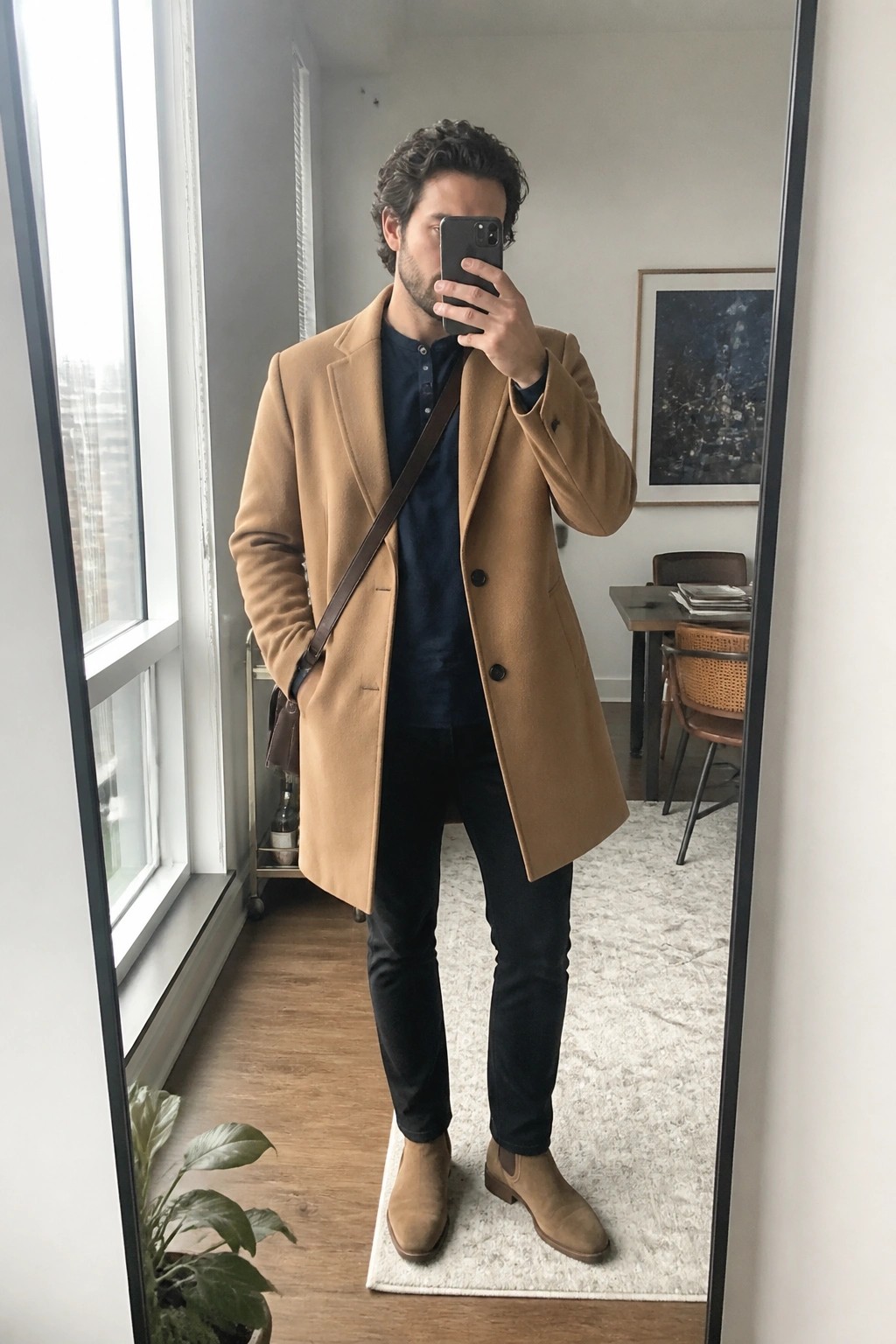 Man in camel coat taking mirror selfie in modern room wearing navy shirt.