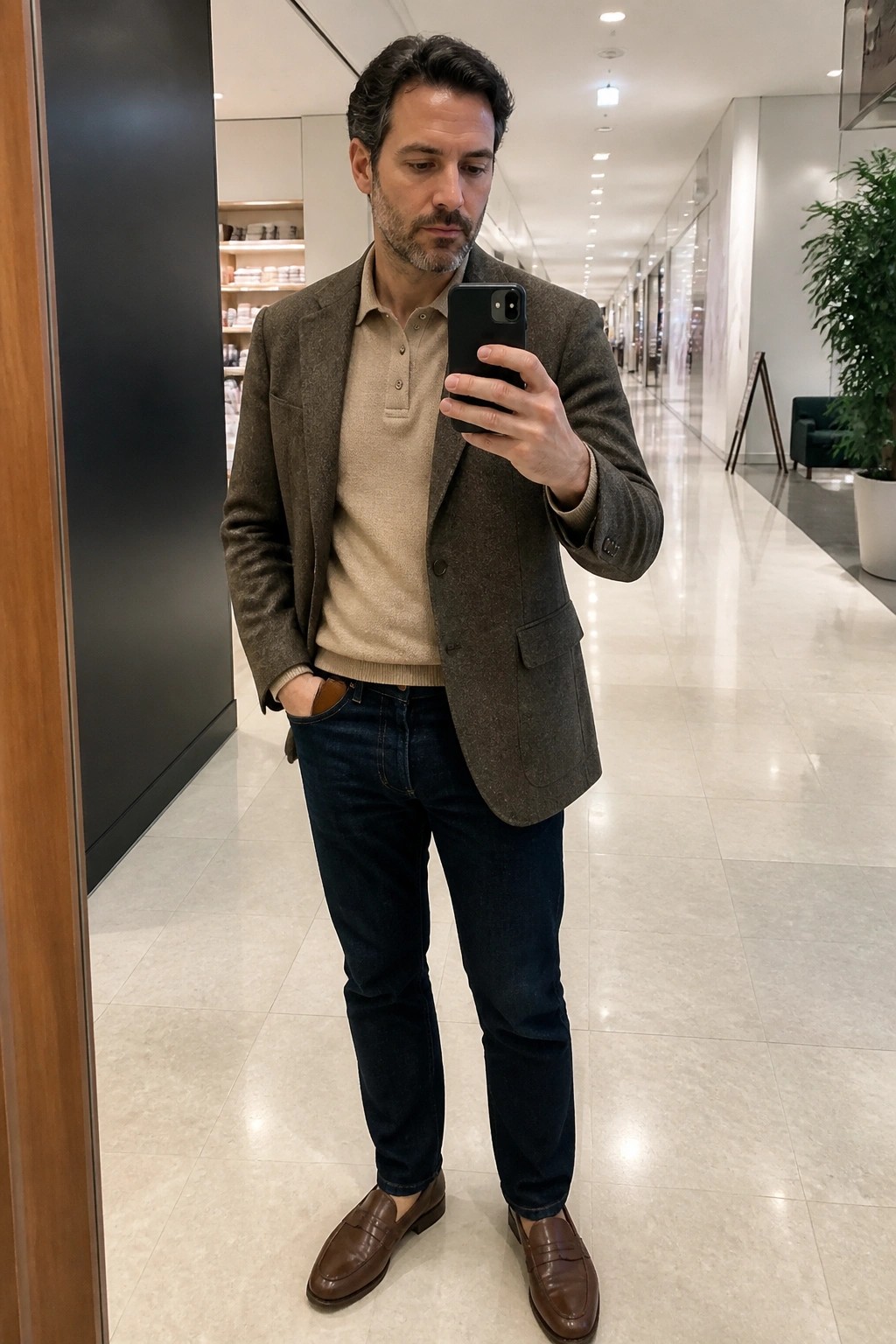 Man wearing brown textured blazer over beige polo sweater, dark blue jeans, and brown leather loafers while holding a phone in a bright indoor hallway.