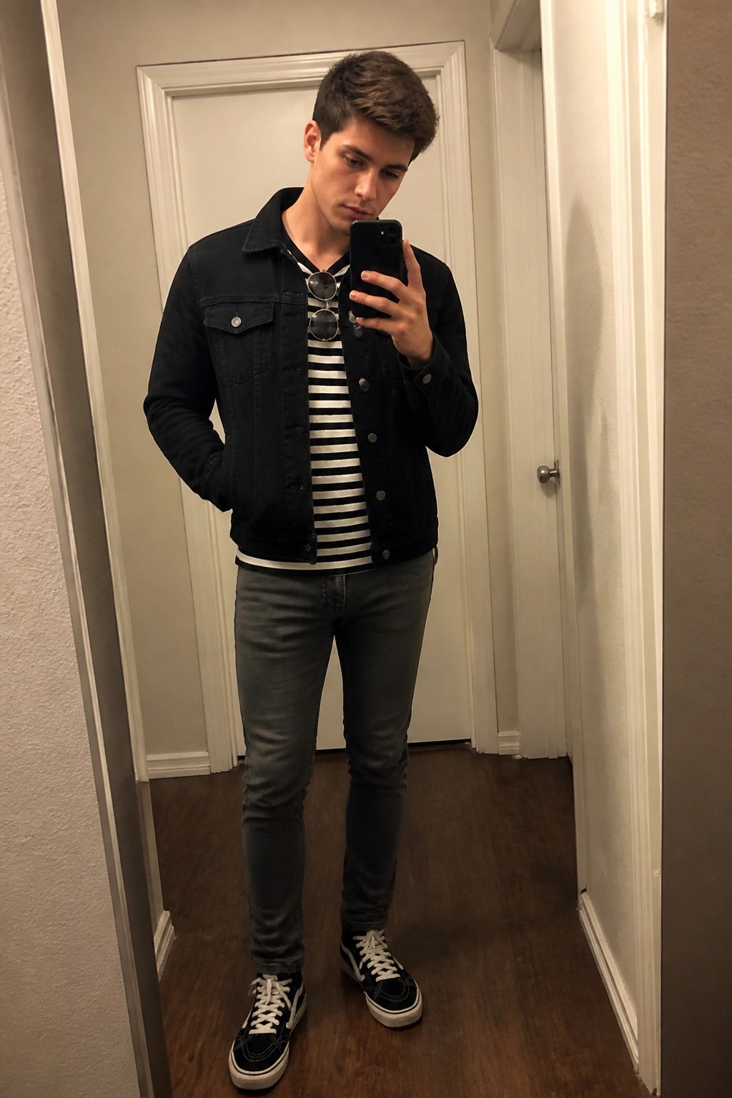 Young man in black denim jacket and striped shirt taking mirror selfie in hallway