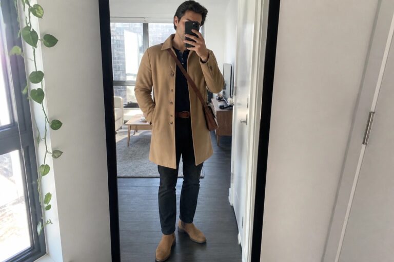 Man in camel coat taking mirror selfie in modern room wearing navy shirt.