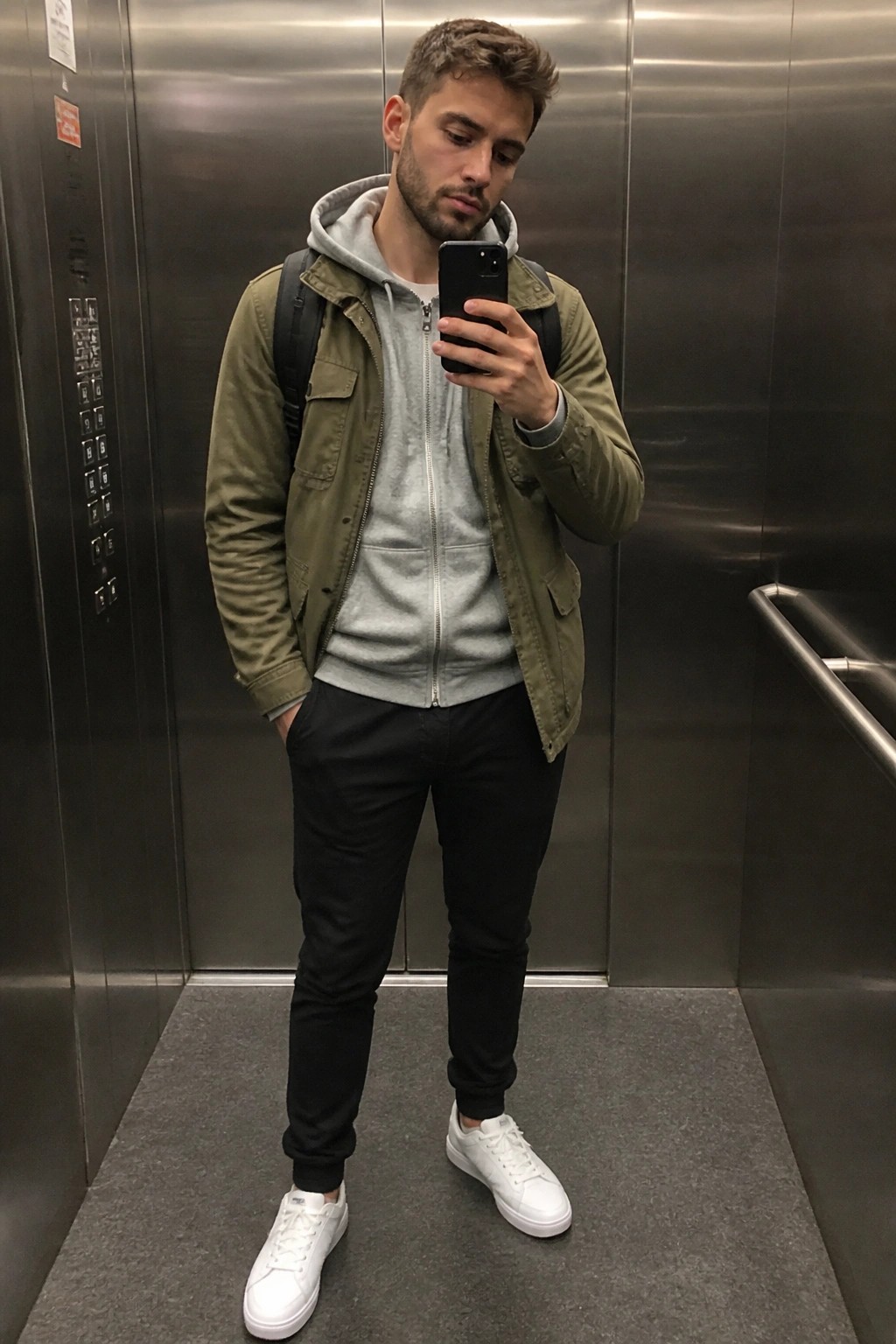 Man in olive jacket over gray hoodie taking selfie in elevator with backpack