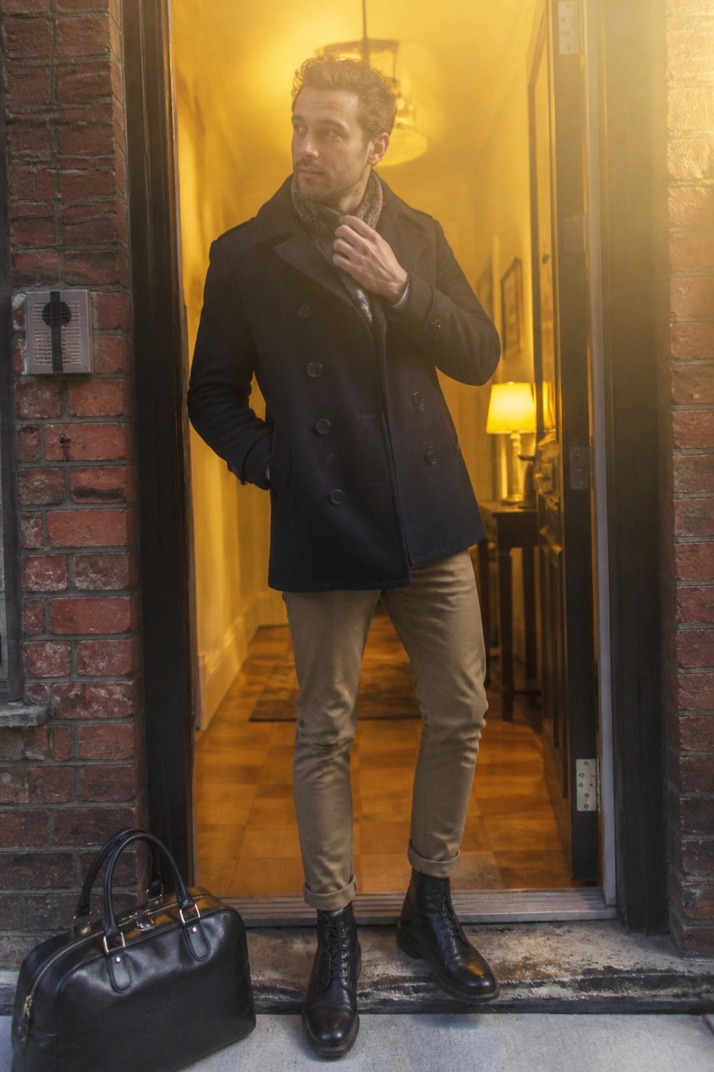 Man in navy peacoat adjusts scarf beside black leather bag in brick doorway