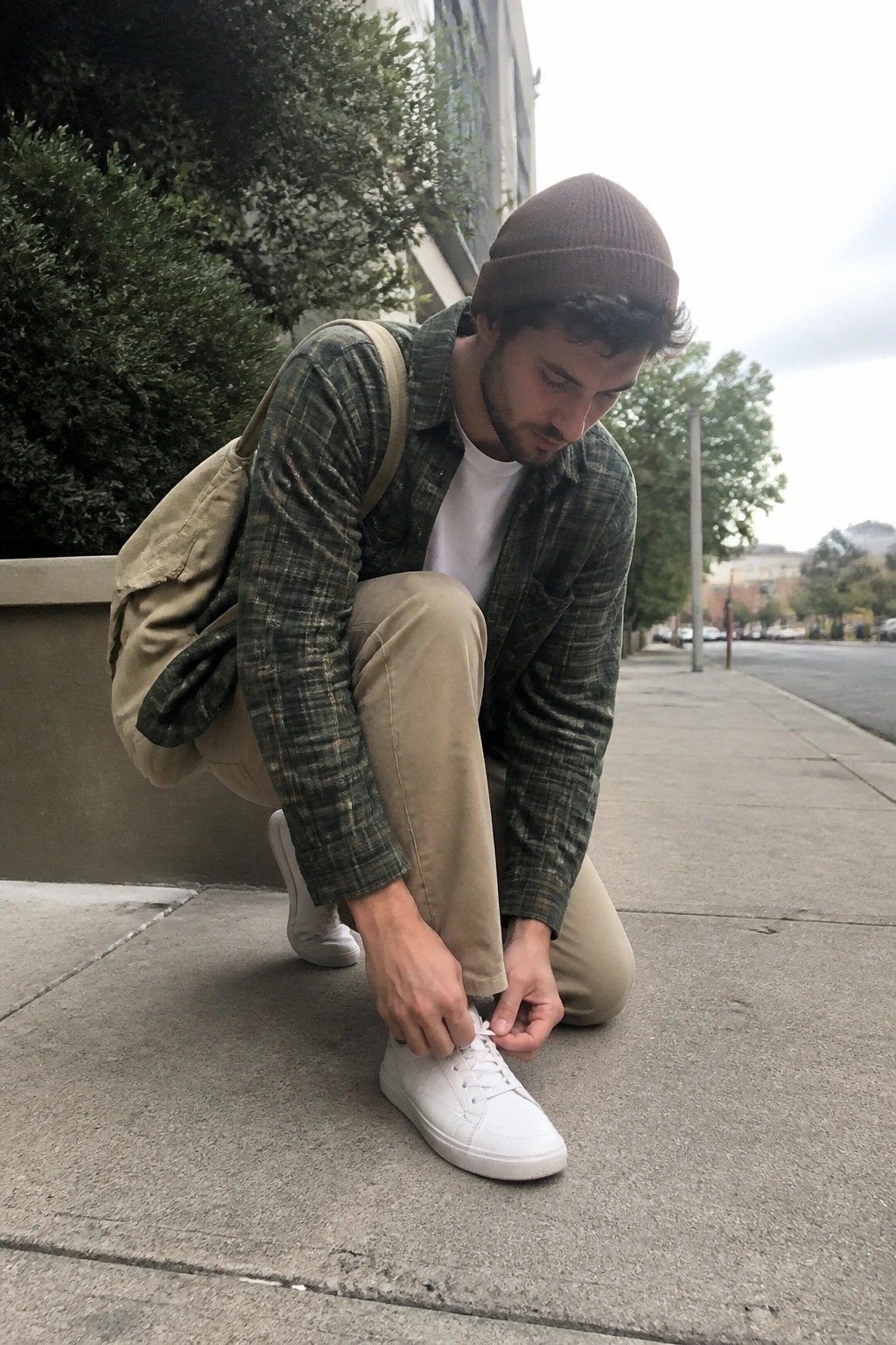 Man in plaid shirt and beanie tying white sneaker on city sidewalk