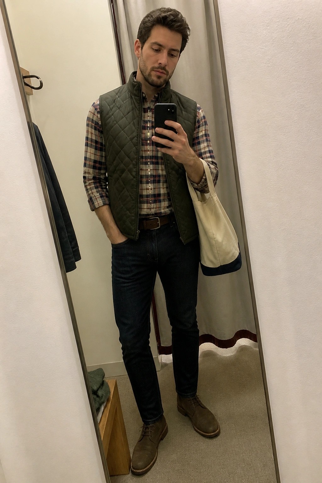 Man in green quilted vest, plaid shirt, dark jeans, brown boots holding tote bag