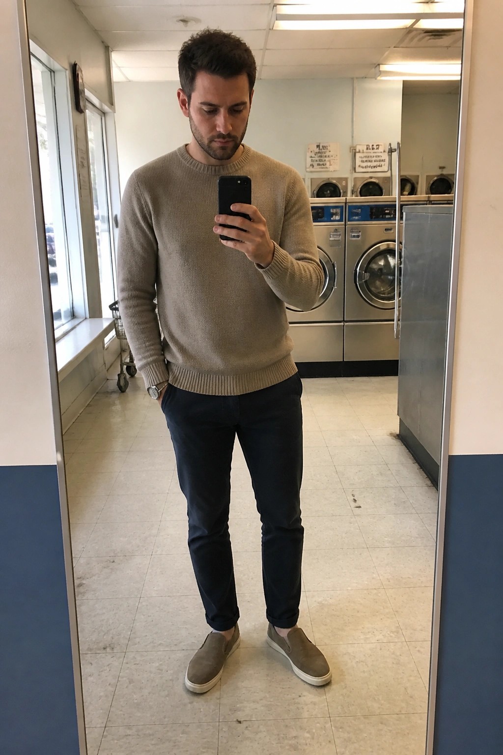 Man in beige sweater taking mirror selfie in laundromat with washing machines behind