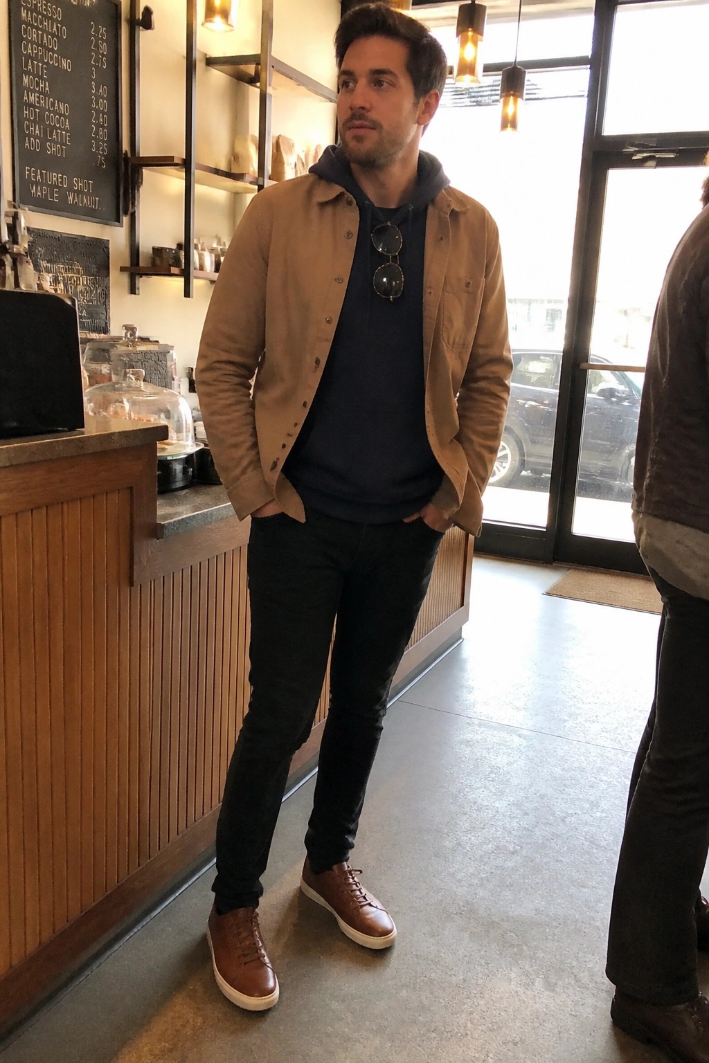 Man in tan jacket over navy hoodie stands in modern coffee shop interior