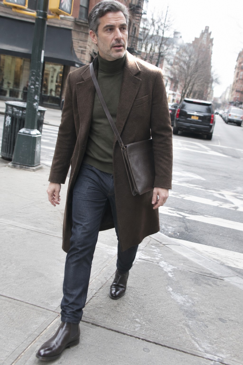 A man wearing a long brown overcoat, olive turtleneck, dark jeans, brown ankle boots, and a dark leather crossbody bag.