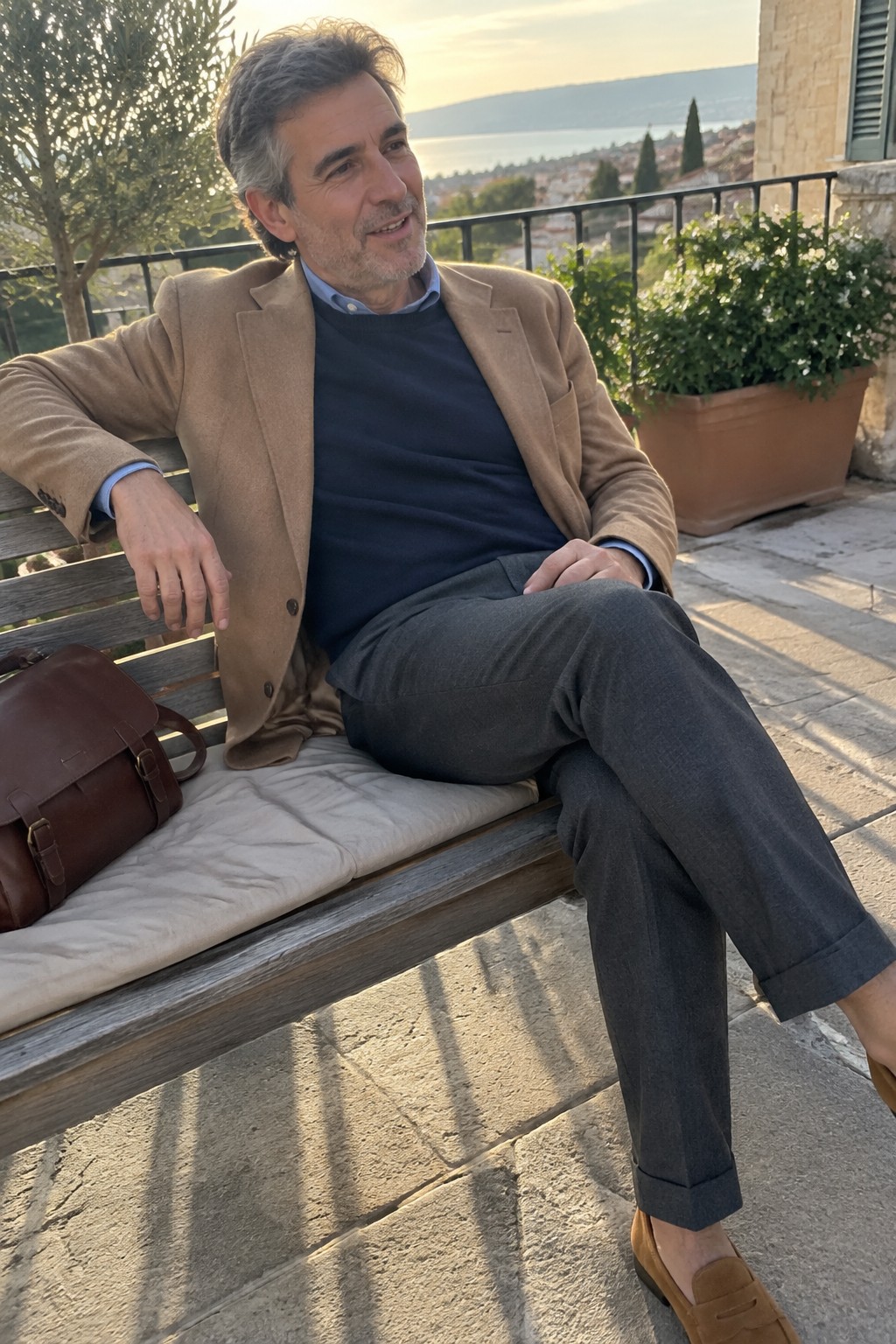 A man in a camel blazer, navy sweater, blue shirt, gray cuffed trousers, and brown suede loafers sits on a bench.