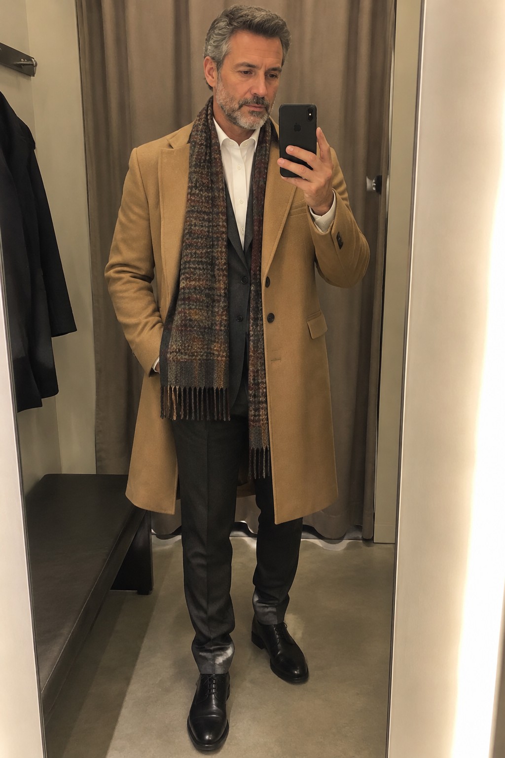 A man wearing a camel overcoat over a charcoal suit and white shirt, with a brown patterned scarf and black leather dress shoes.
