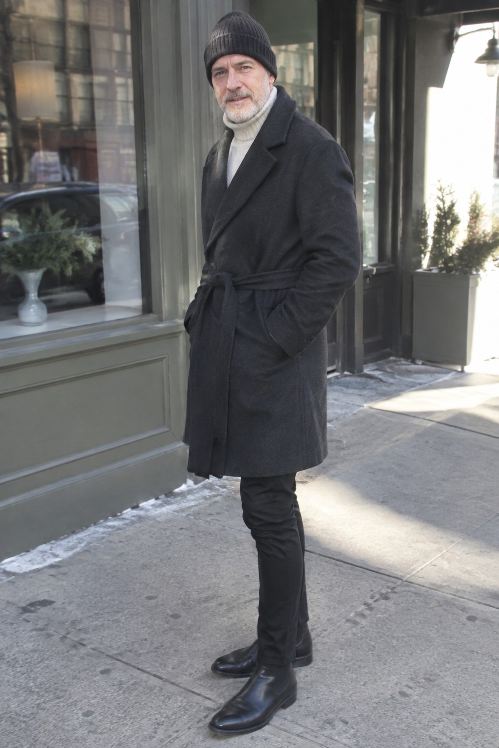 Man over 50 wearing a charcoal belted wool overcoat, cream ribbed turtleneck, black trousers, black Chelsea boots, and a dark knit beanie.