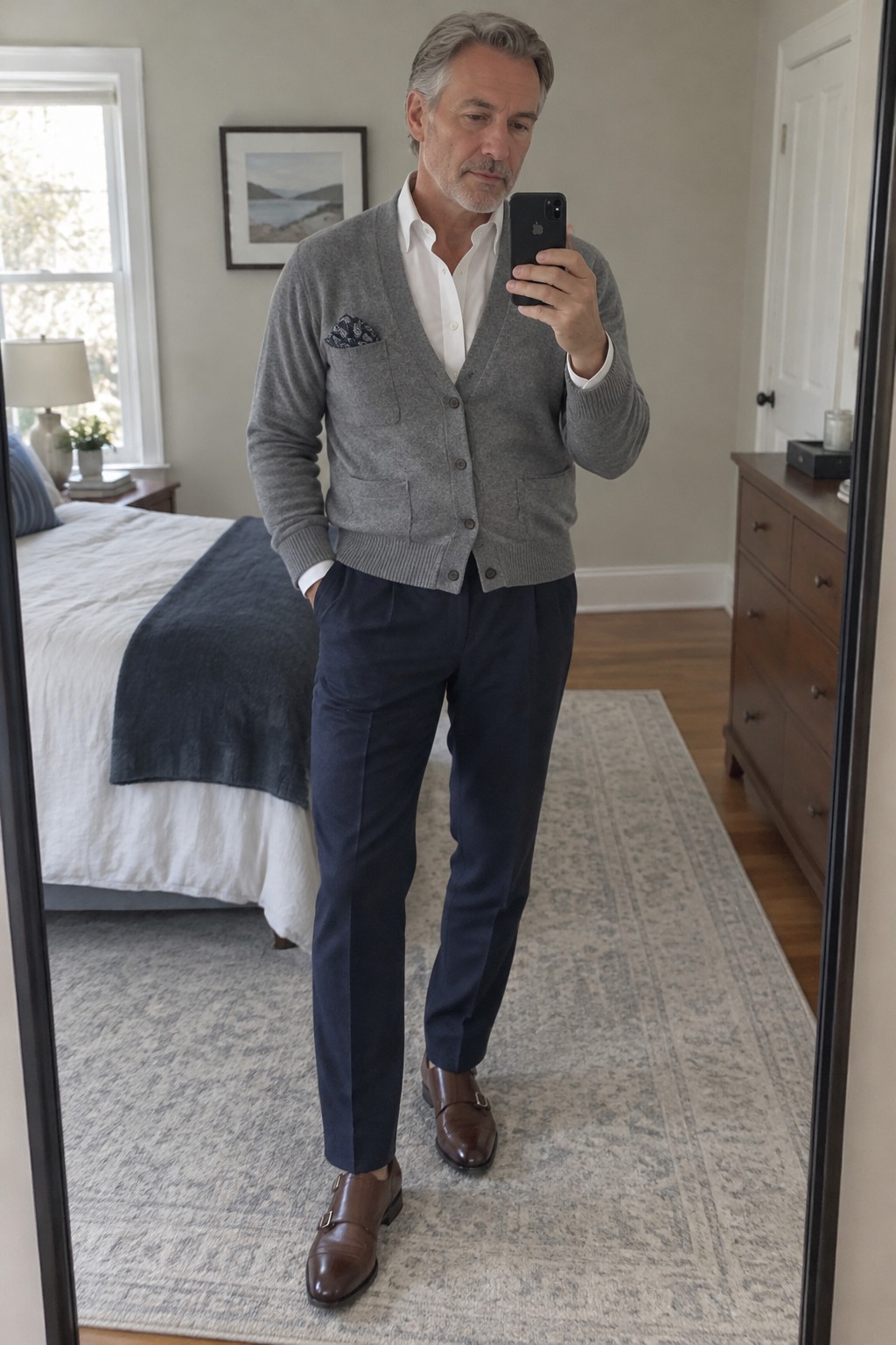 A man wearing a gray cardigan over a white shirt with a patterned pocket square, navy trousers, and brown double-monk shoes.