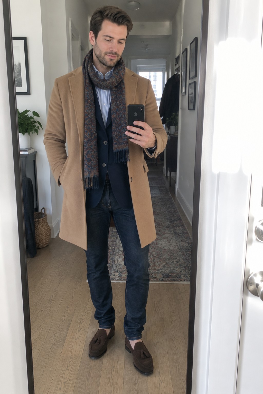 A man wearing a camel overcoat over a navy blazer and blue striped shirt, with a patterned scarf, dark jeans, and brown tassel loafers.