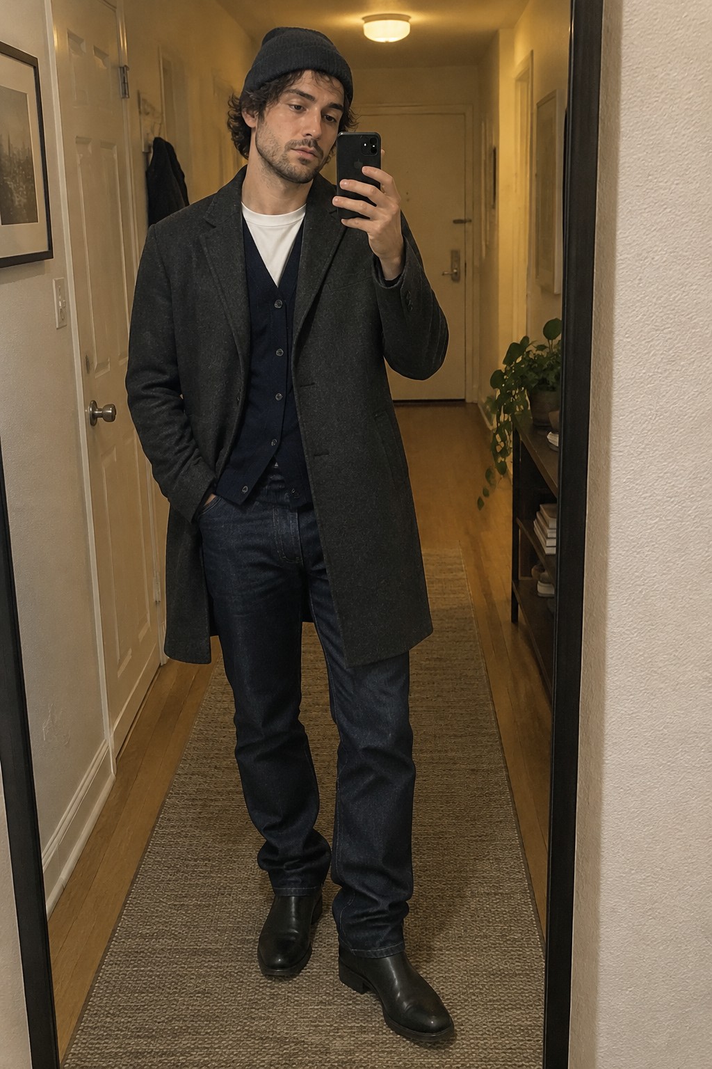 Man wearing a long charcoal coat over a navy waistcoat and white T-shirt, with dark jeans, black Chelsea boots, and a dark knit beanie.