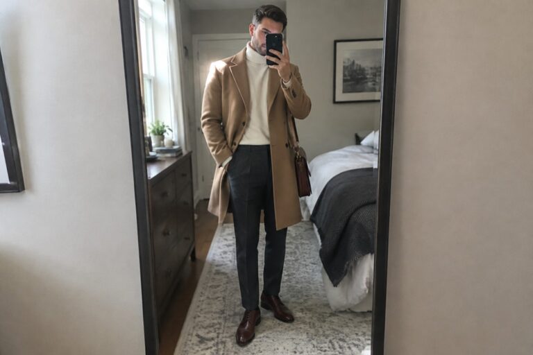 17 Polished Semi-Formal Old Money Winter Outfits for Men Styled Around Tailored Coats A man wears a camel overcoat over a cream turtleneck with charcoal trousers, brown leather lace-up shoes, and a dark brown shoulder bag.