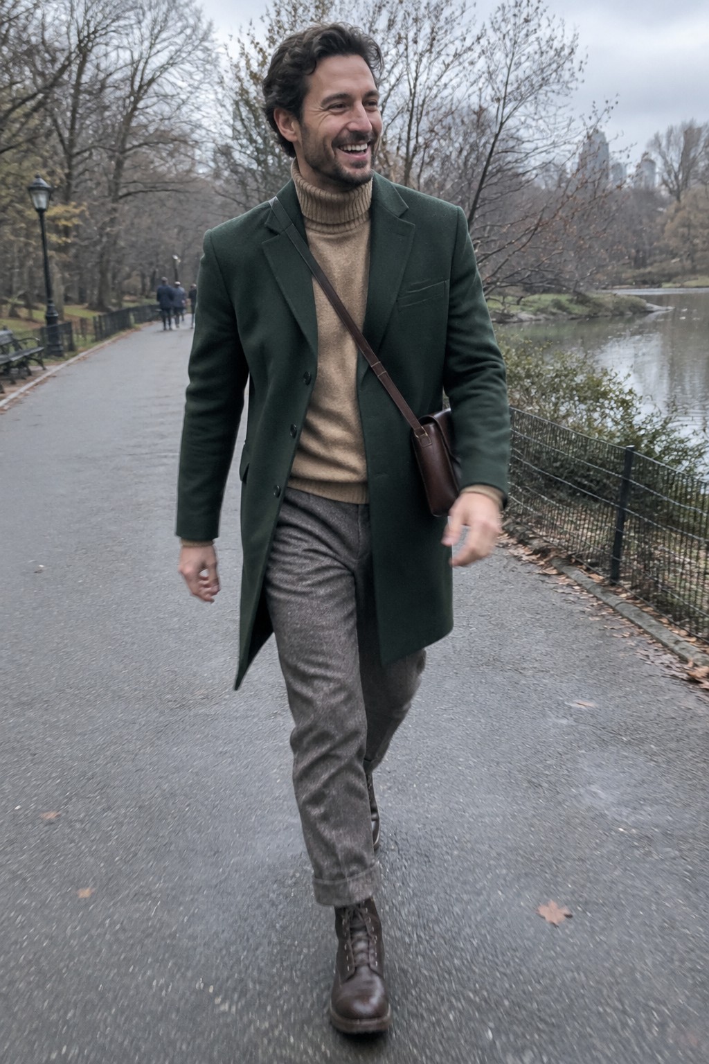 A man wears a dark green tailored coat over a tan ribbed turtleneck, gray tweed trousers, brown leather boots, and a brown crossbody bag.
