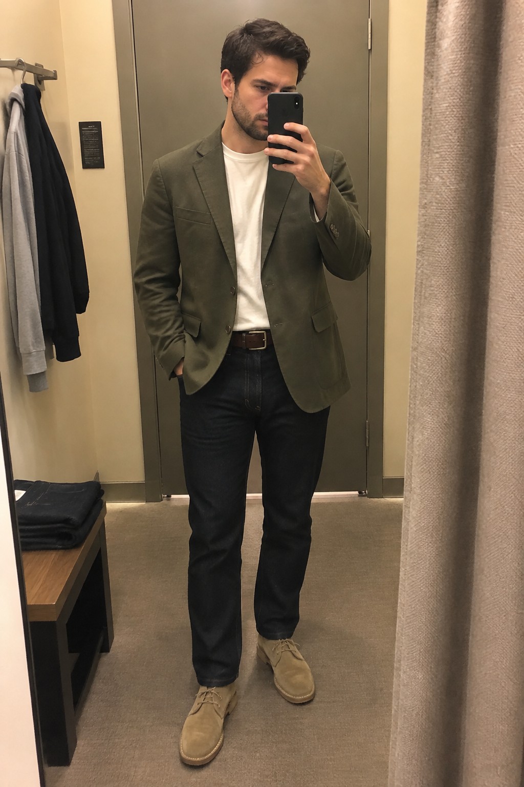 A man wears an olive blazer over a white crewneck T-shirt with dark straight-leg jeans, a brown leather belt, and tan suede chukka boots.