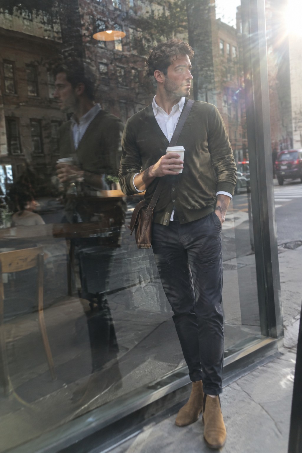 Man wearing a dark olive cardigan over a white button-down shirt with dark trousers, brown suede ankle boots, and a brown leather crossbody bag.