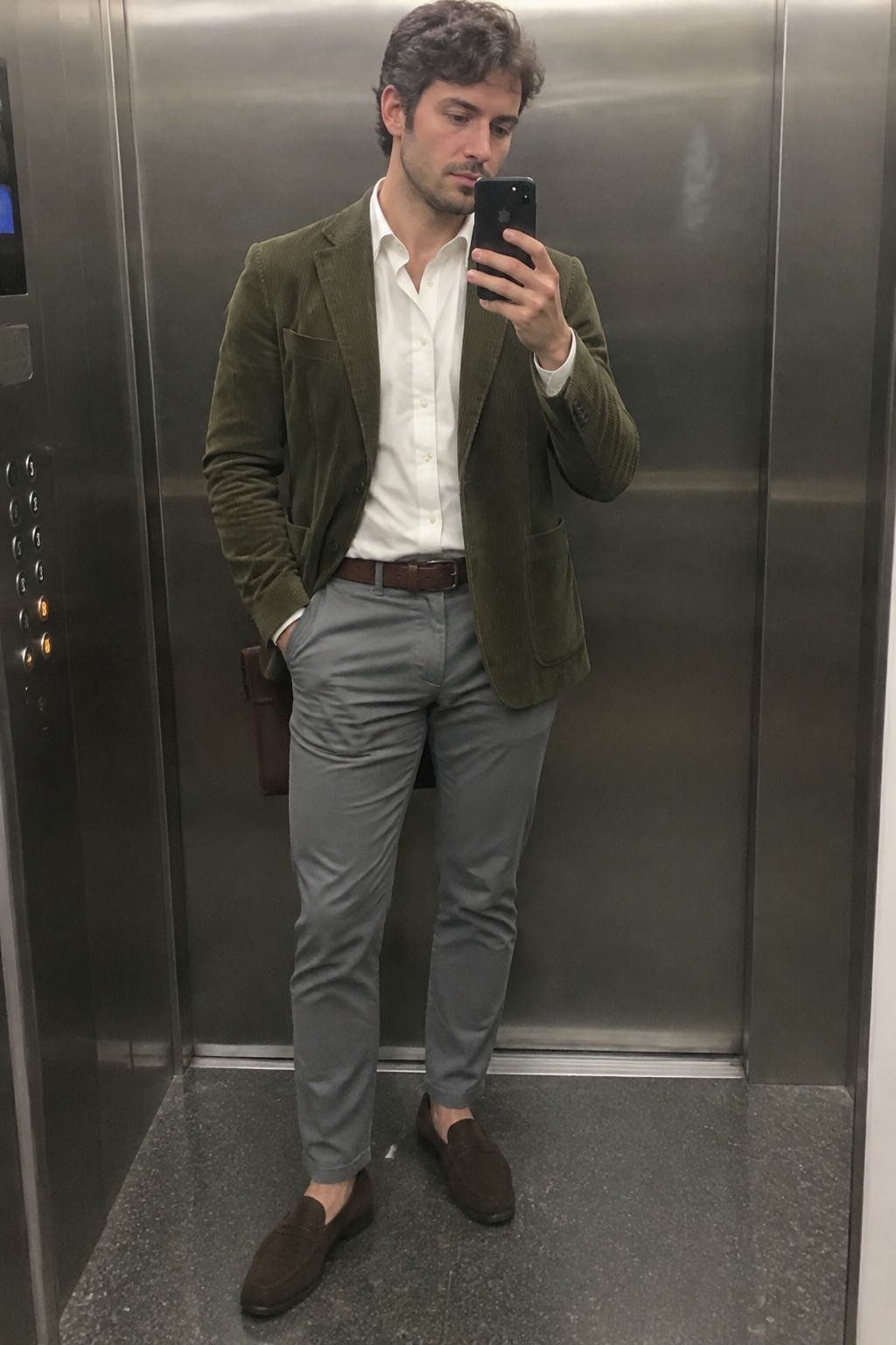 A man wearing an olive corduroy blazer, an open-collar white shirt, gray trousers, a brown belt, and dark brown suede loafers.
