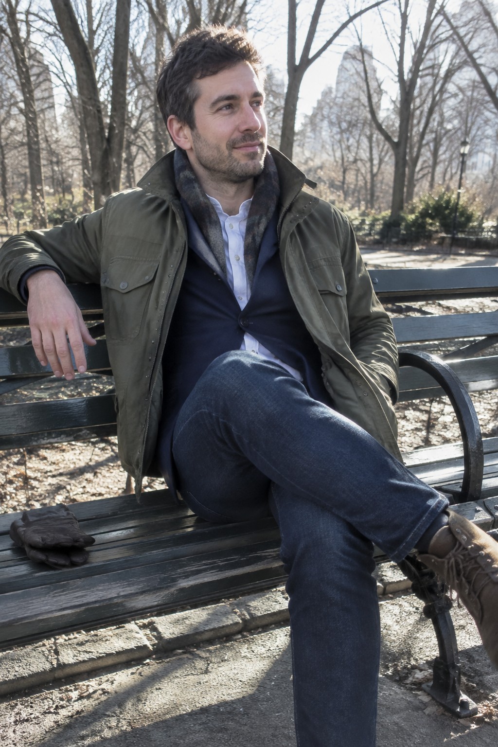 Man wearing an olive field jacket over a navy cardigan, white button-down shirt, dark jeans, a muted scarf, and brown lace-up boots while seated on a bench.