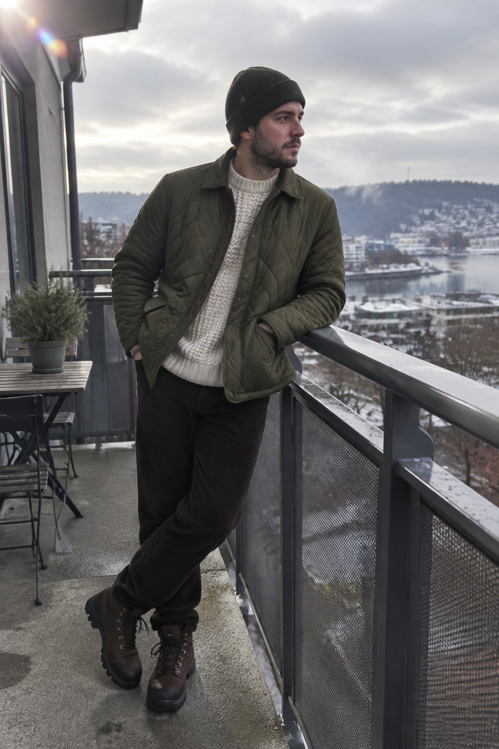 A man wearing a dark olive quilted jacket over a cream cable-knit sweater, dark brown corduroy trousers, a black beanie, and brown lace-up boots.