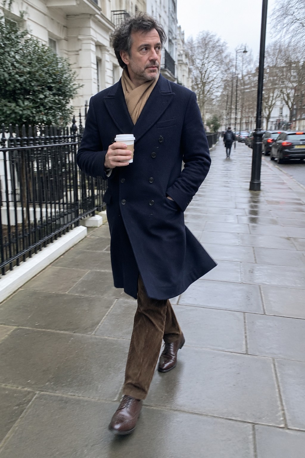A man wears a navy double-breasted overcoat, camel scarf, brown corduroy trousers, and brown leather lace-up shoes while holding a takeaway coffee cup.